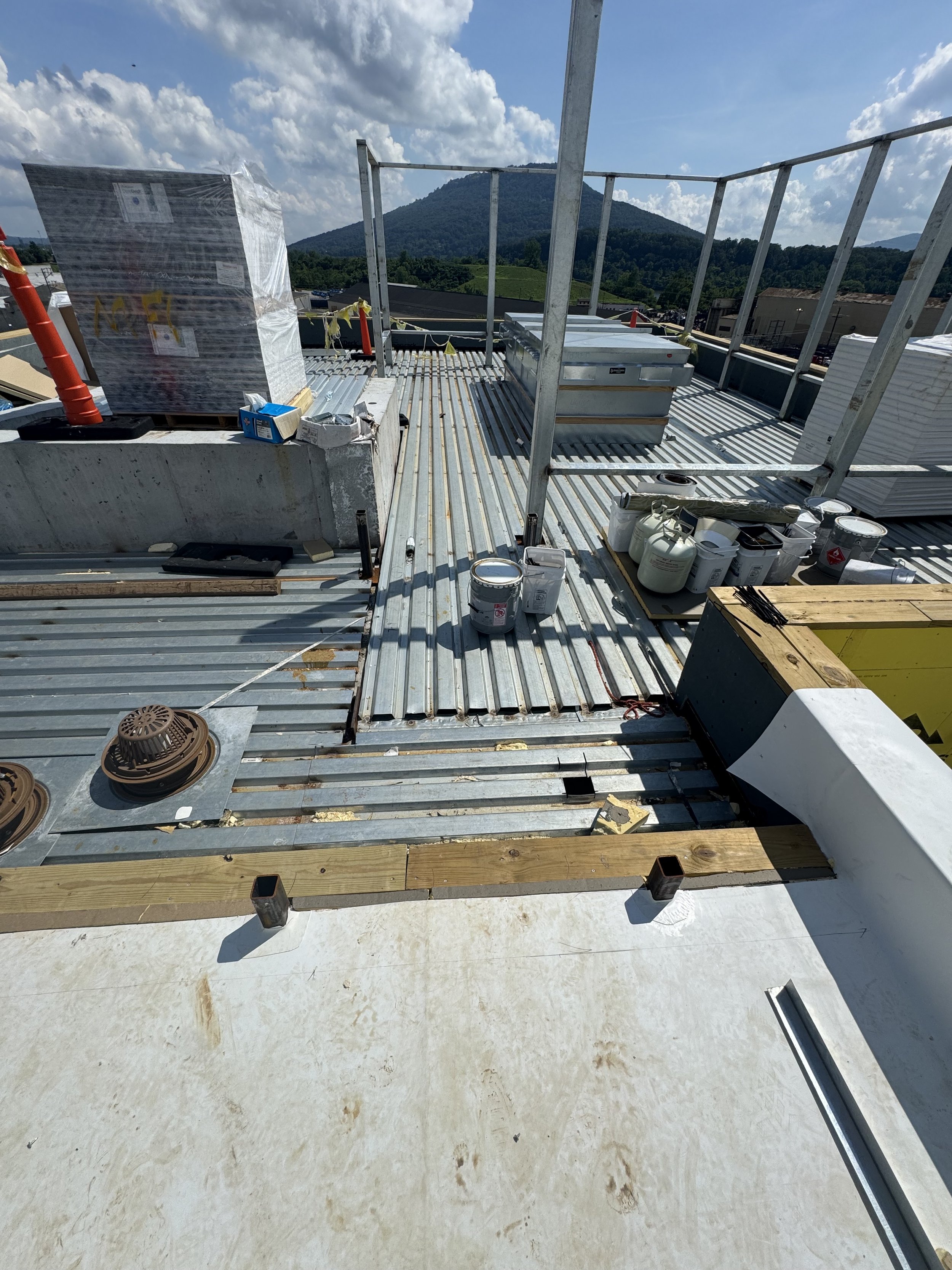 Construction site on a rooftop with metal decking, construction materials, and tools, with a mountain and blue sky with clouds in the background.
