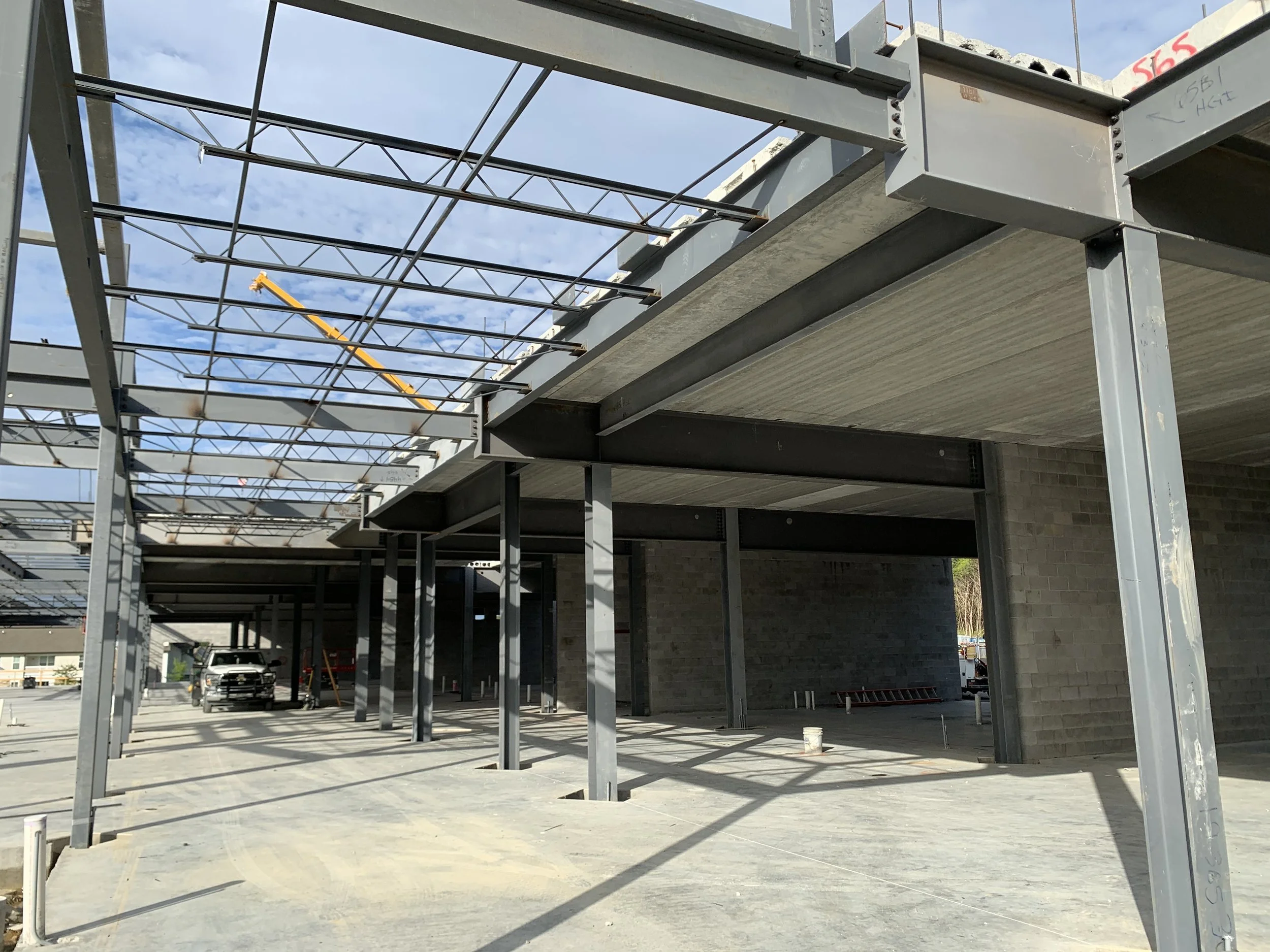 Construction site with steel framework for a building, with parked trucks and construction equipment, and a blue sky with scattered clouds.