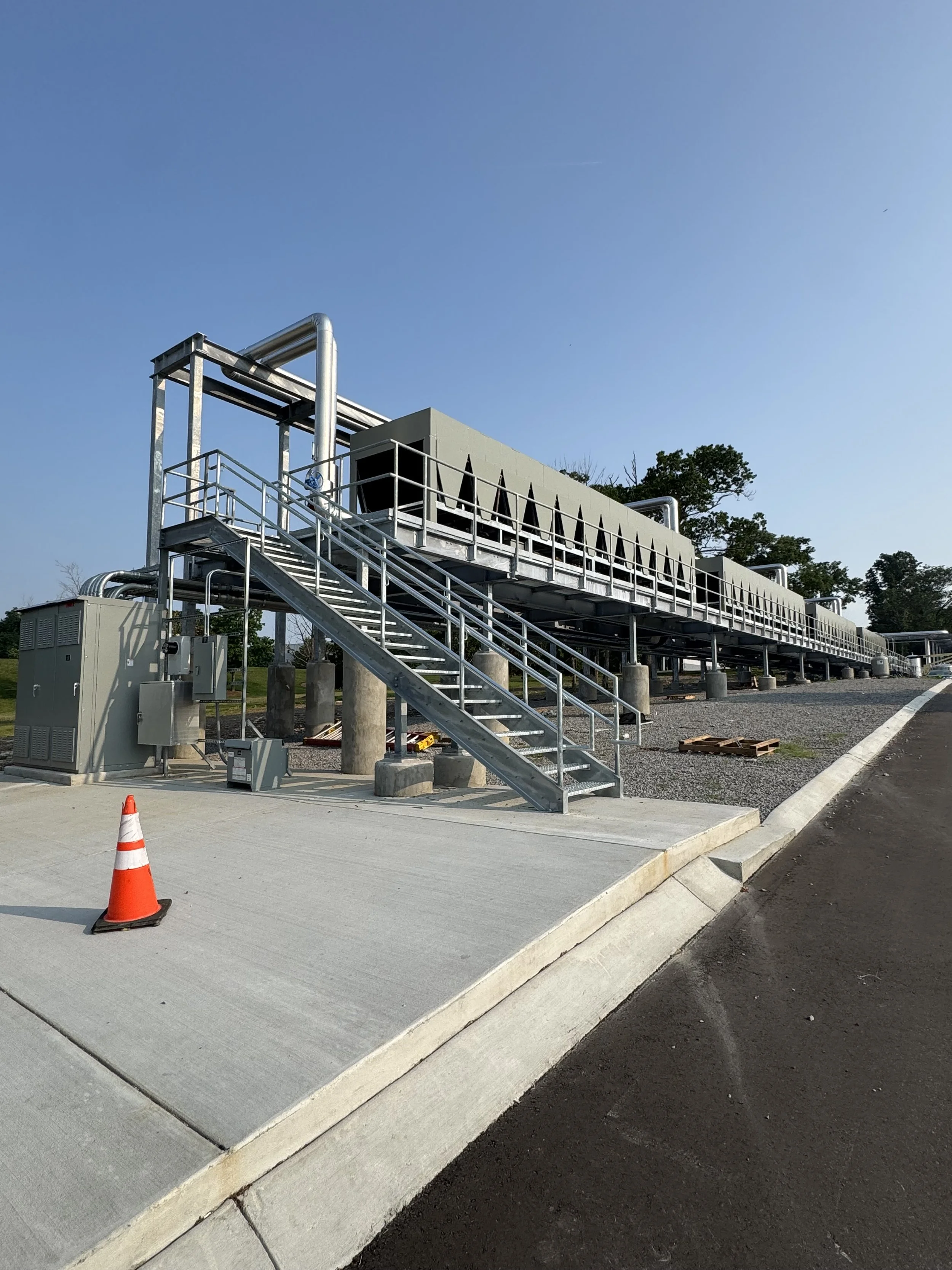 Industrial facility with metal pipes and stairs, situated outdoors on a concrete platform with a traffic cone nearby.