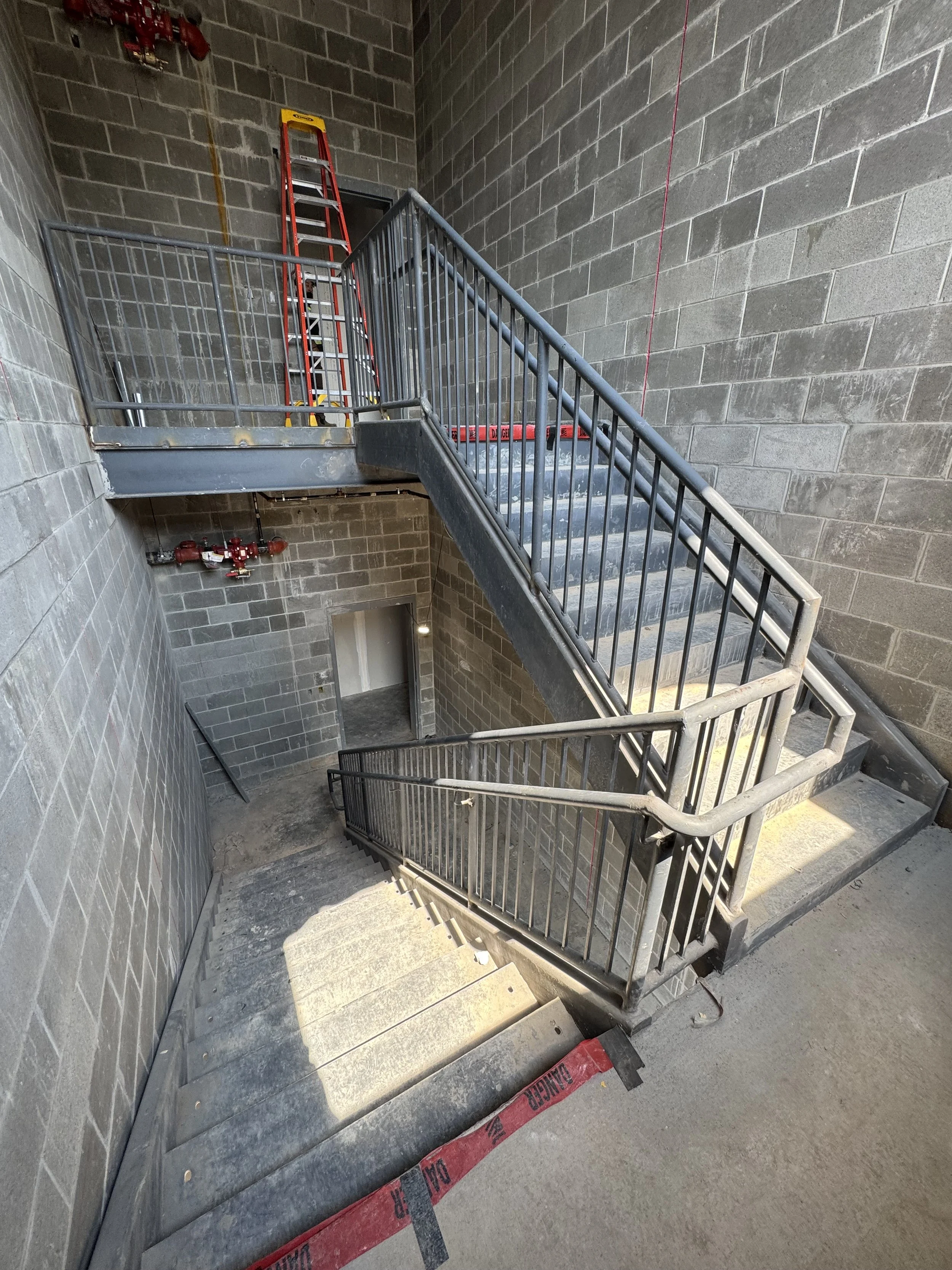 A stairwell inside a building under construction, with concrete steps, metal railings, exposed gray brick walls, a red ladder, and construction warning tape on the floor.