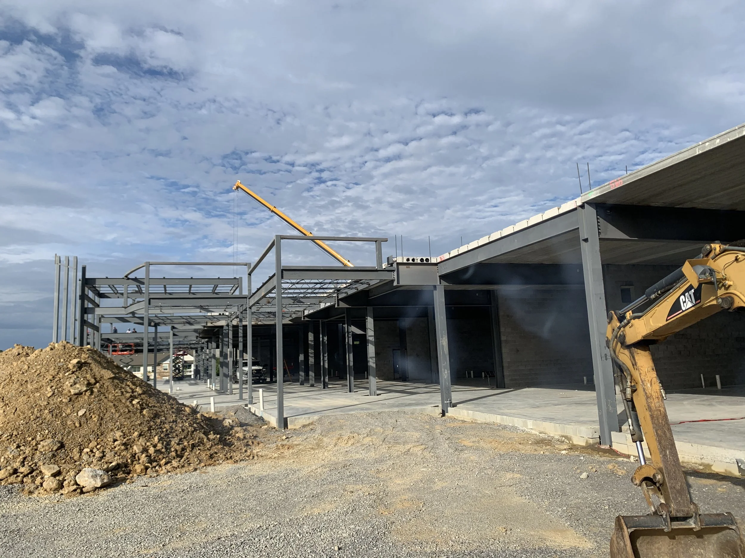 Construction site with steel framework for a building, a pile of dirt in the foreground, a yellow excavator on the right, and a crane in the background under a cloudy sky.