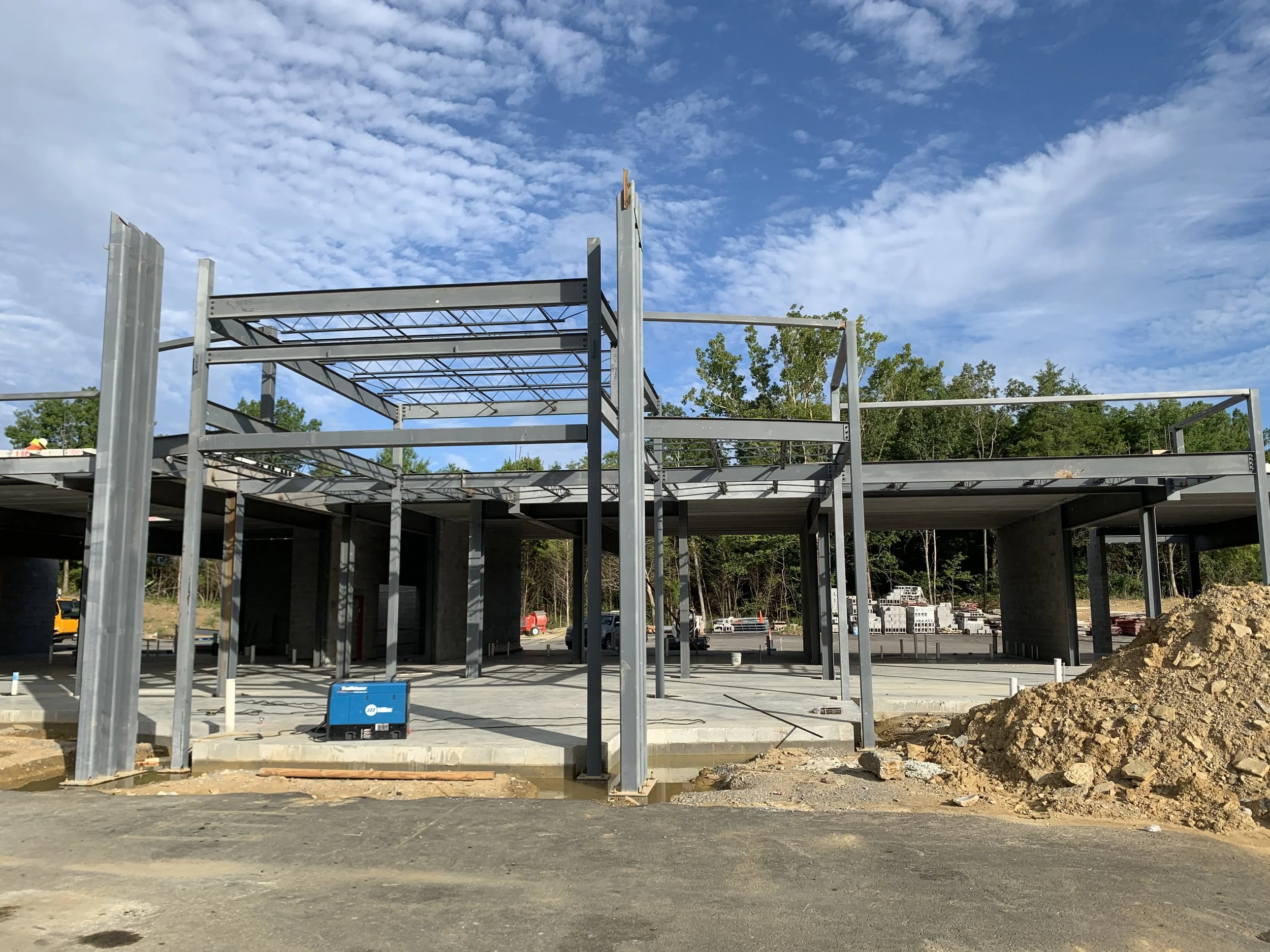Construction site with steel framework and concrete foundation, under a partly cloudy sky, with construction materials and dirt mounds nearby.