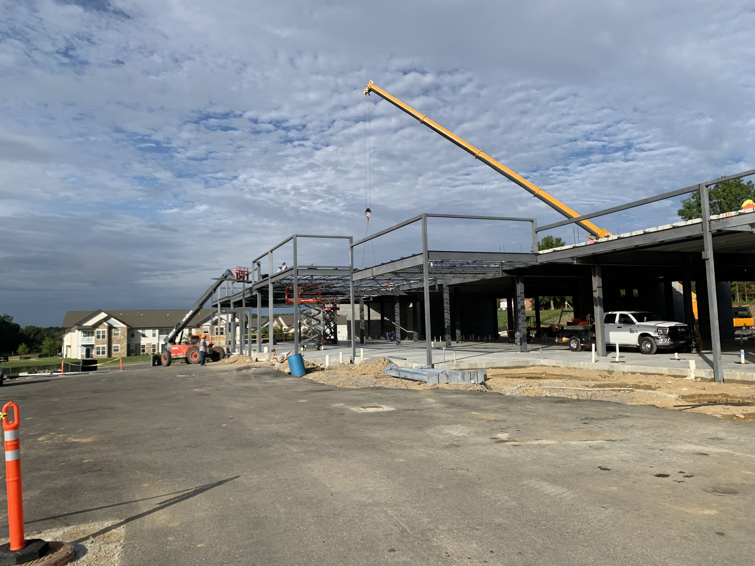 Construction site with a steel framework building under construction, a crane lifting materials, construction vehicles, and a partly paved area under a cloudy sky.