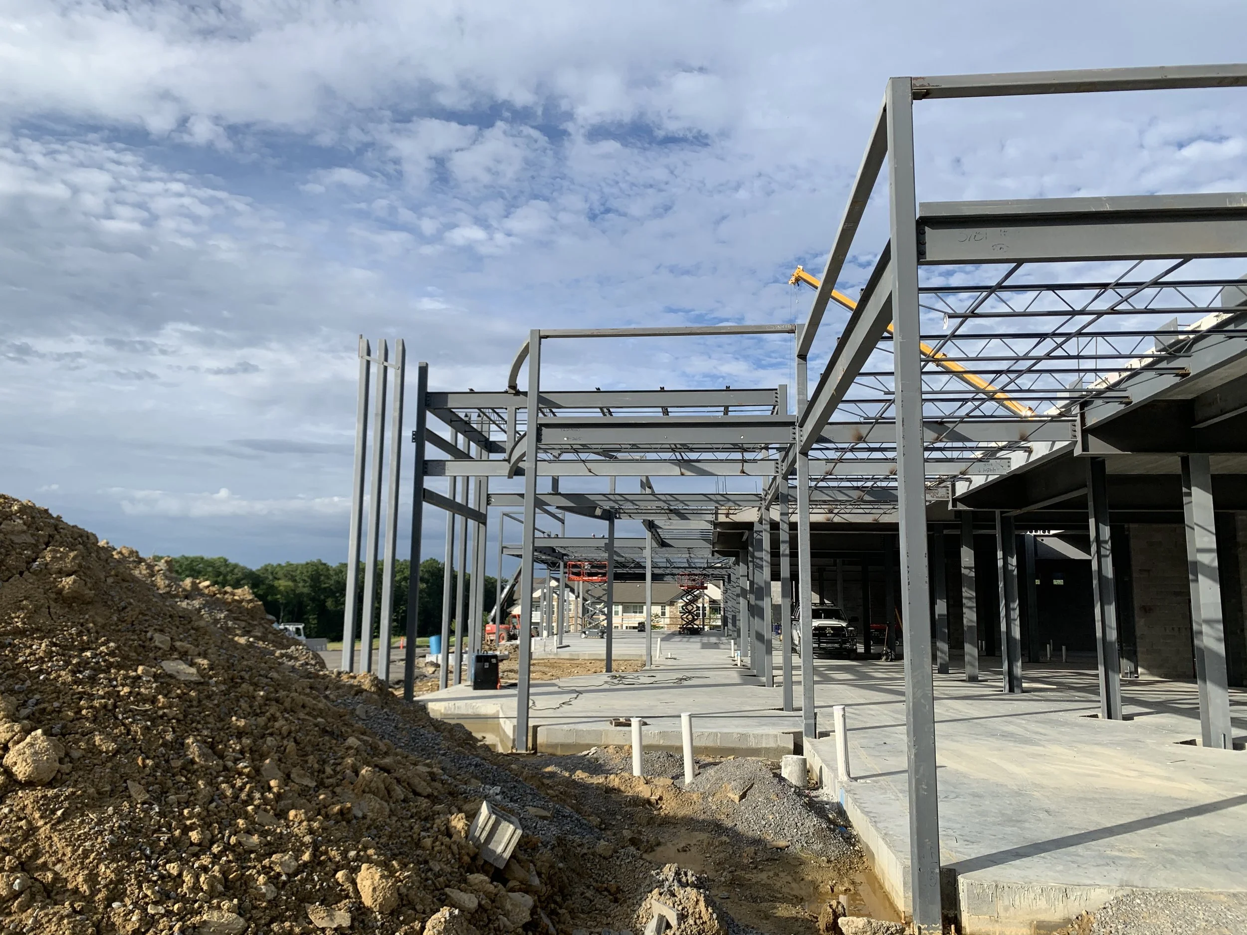 Construction site with steel framework for a building, gravel on the ground, and a crane in the background under a partly cloudy sky.