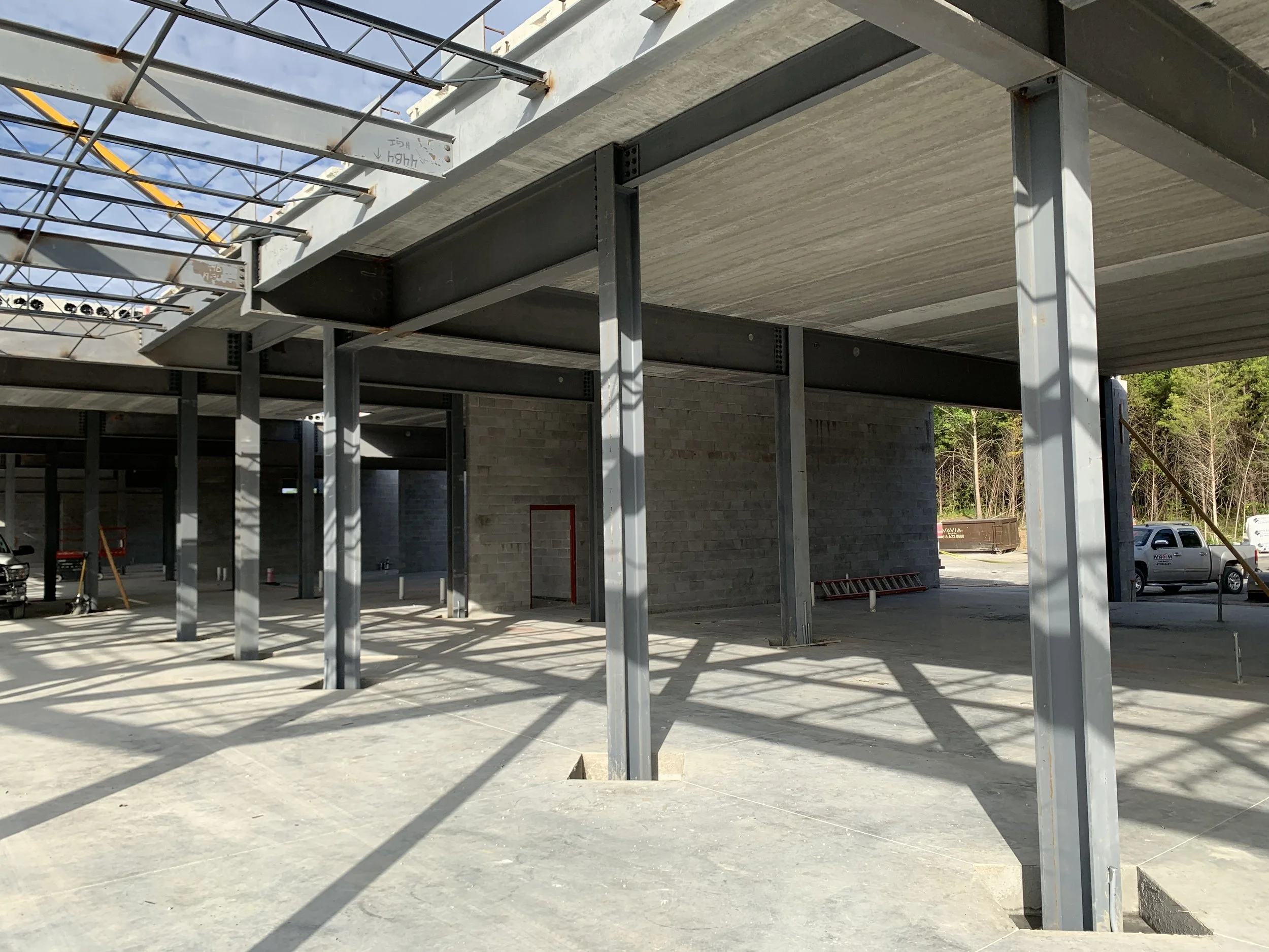 Construction site of a building with steel beams, concrete floors, and brick walls, with vehicles parked outside and trees in the background.
