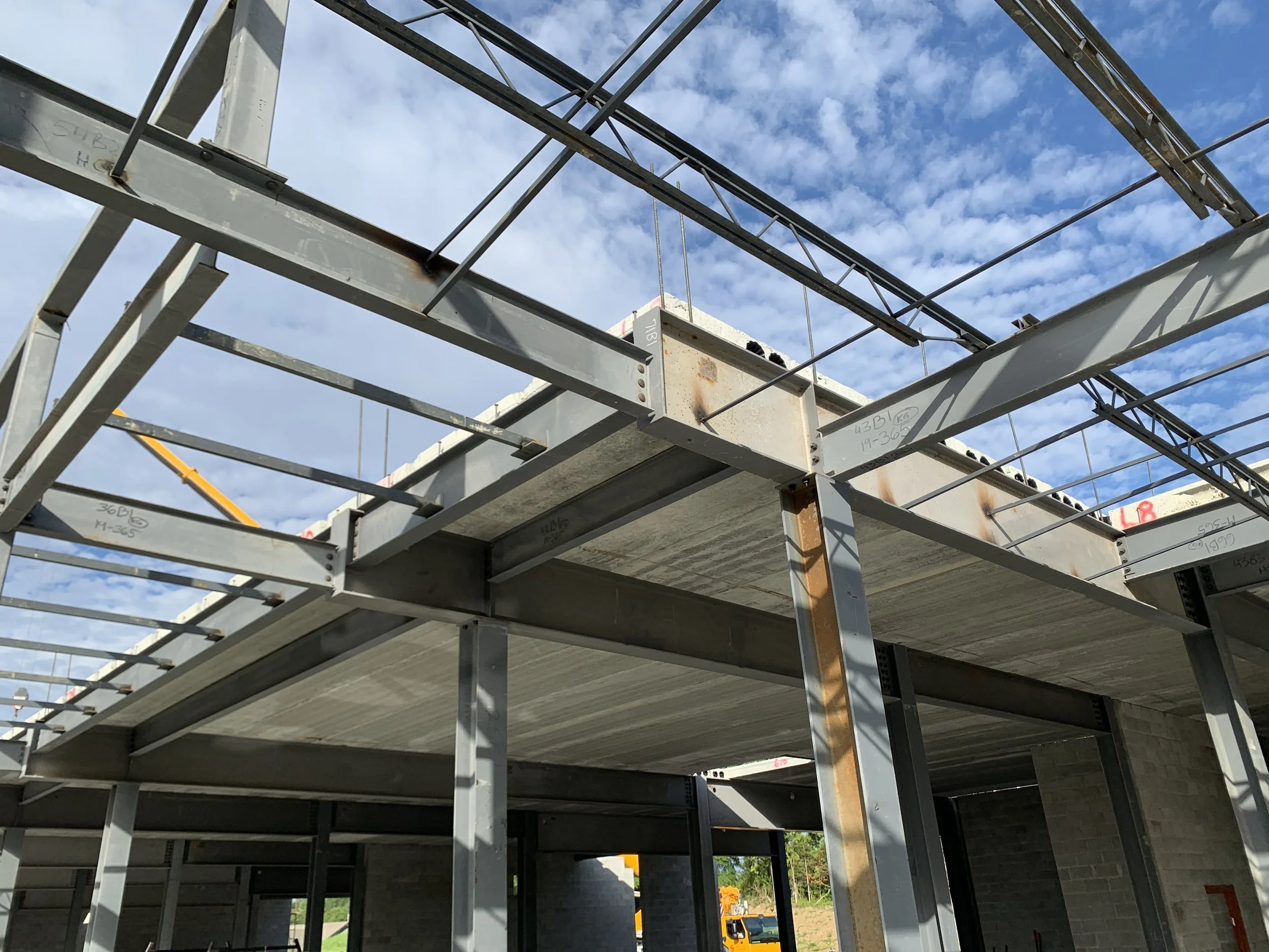Construction site showing steel framework and concrete floors of a building under construction, with a blue sky and scattered clouds in the background.