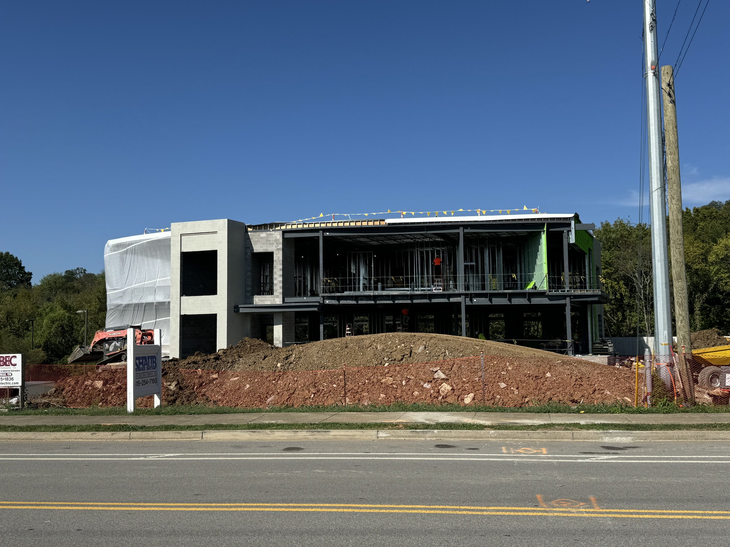 Under construction building with exposed steel framework, concrete walls, and a multi-level structure, set behind a mound of dirt along a road.