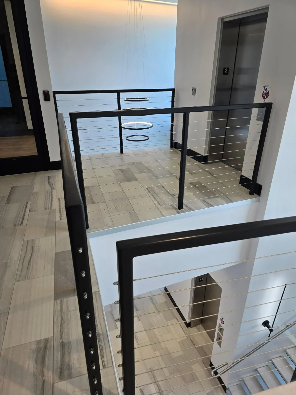 Top view of an indoor staircase and elevator area with black and metal railings, tiled floor, and white walls, with elevator doors on the right and a light fixture on the ceiling.
