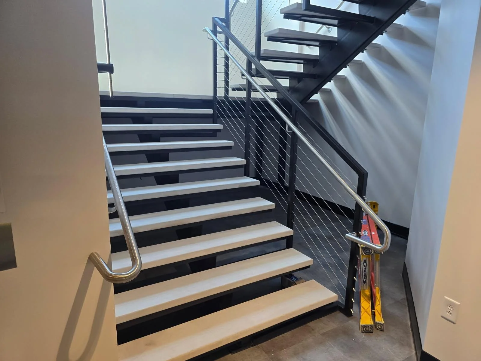 Interior view of a modern staircase with black framing, white steps, and metal railings, partially built with a ladder nearby.