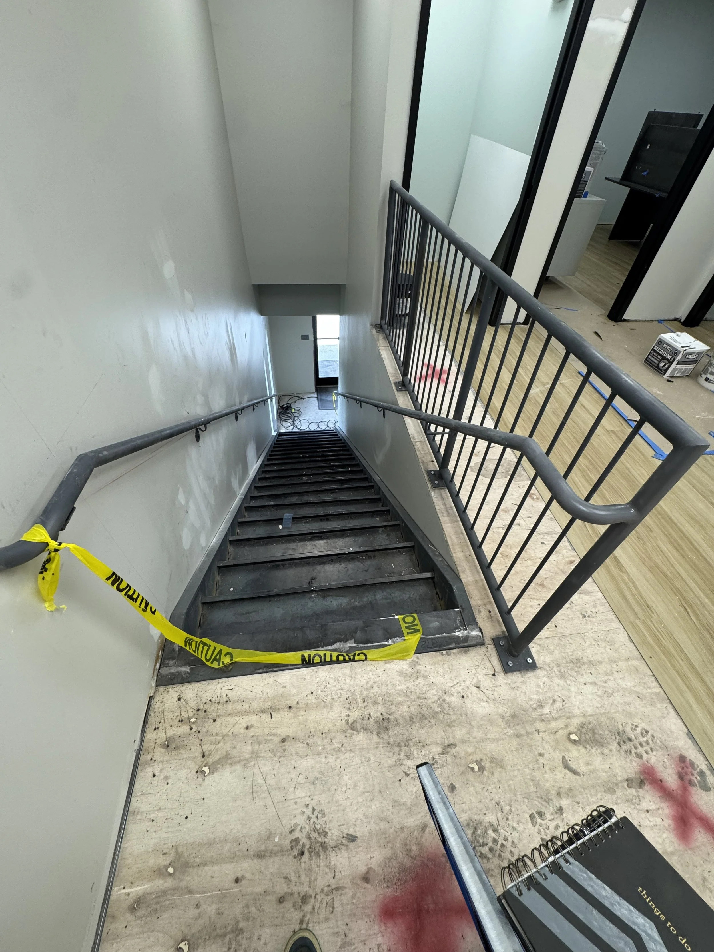 Staircase leading down to a lower floor under construction, with caution tape across the top, drywall and construction materials nearby, and an office space visible at the top.