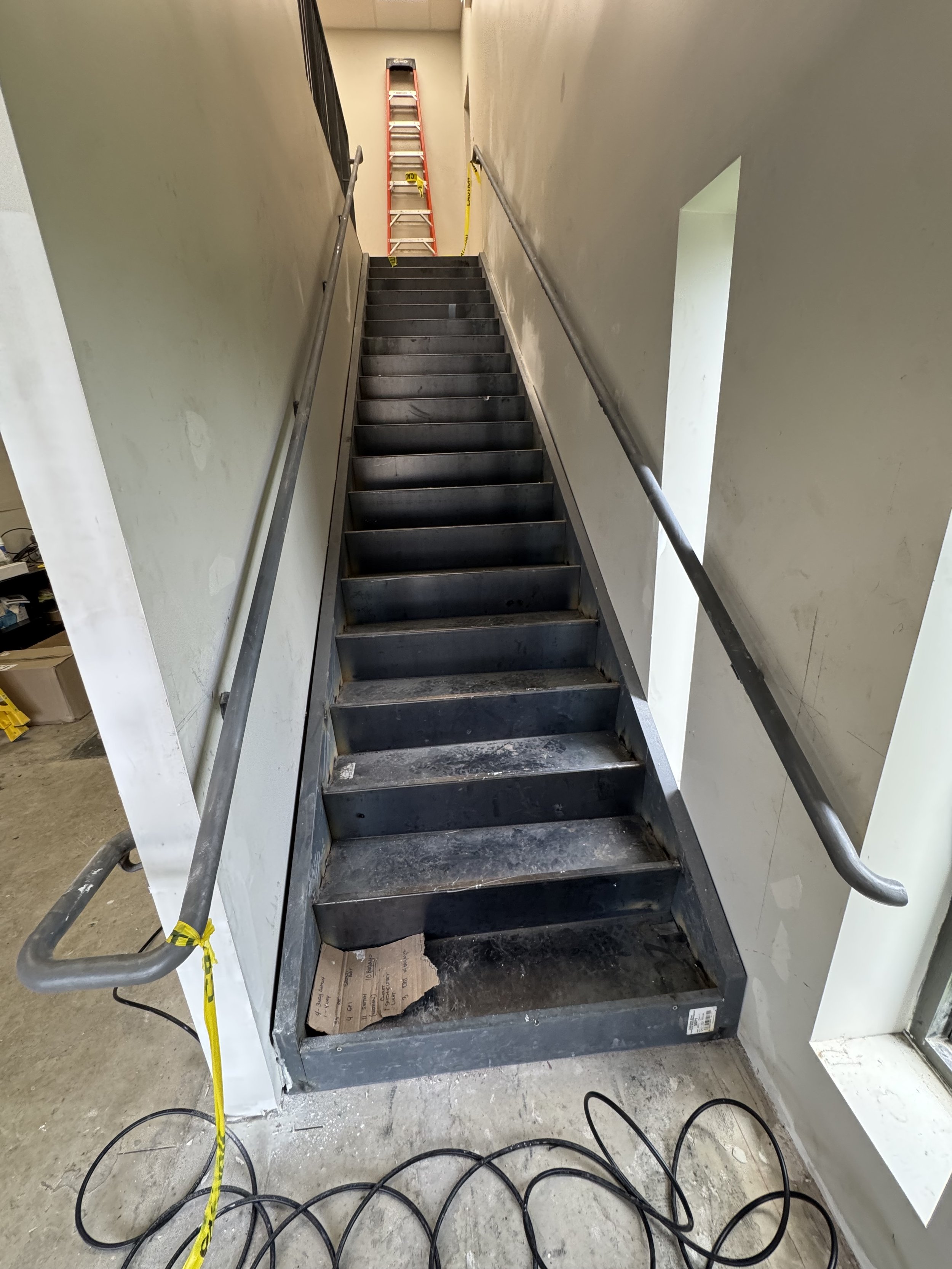Steel staircase with black steps, handrails on both sides, and a ladder at the top. Yellow caution tape is on the wall near the ladder, and some tools and cords are visible on the floor.