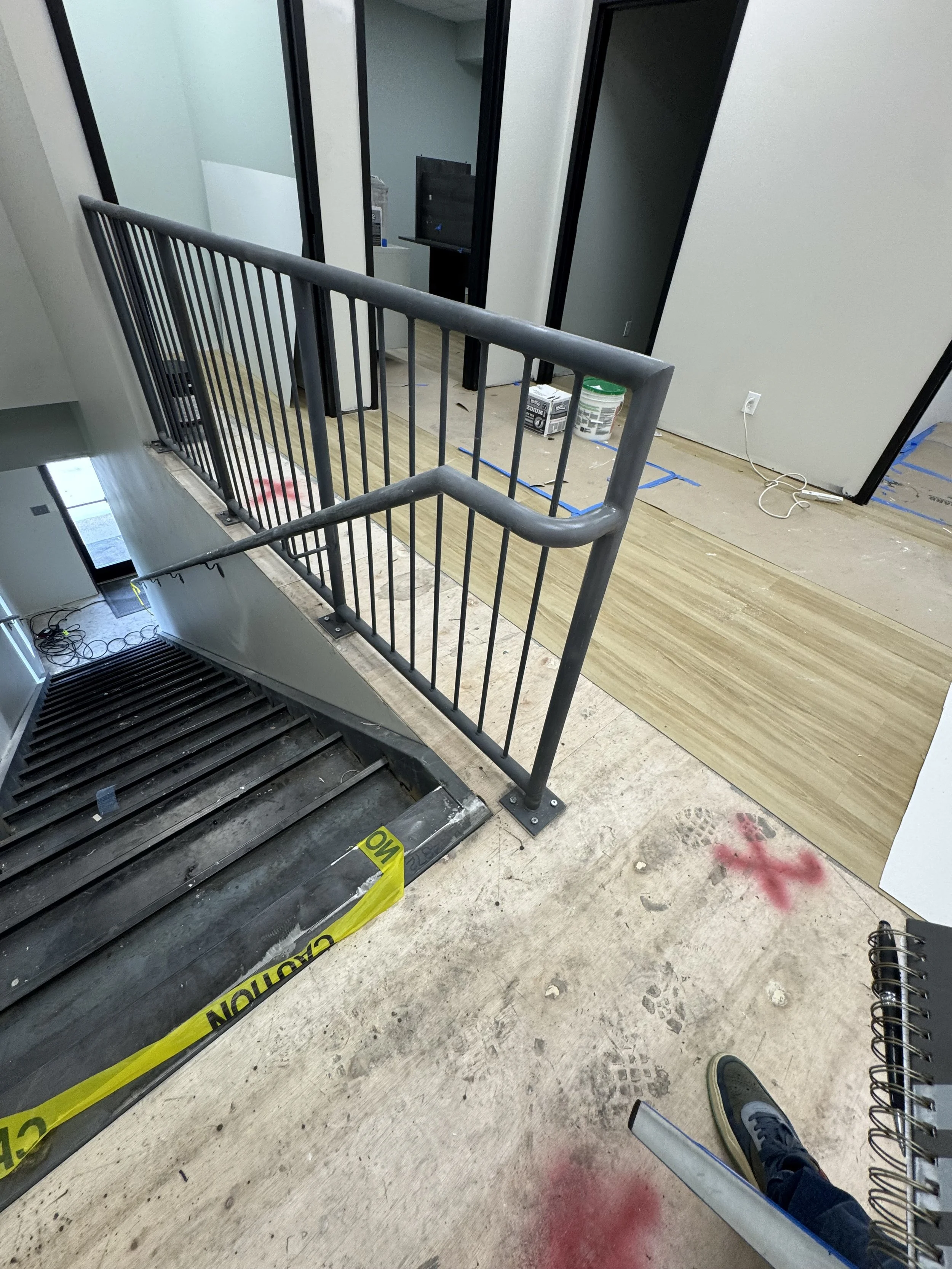 Interior of a building under construction or renovation, showing a staircase with safety railing, unfinished flooring, construction materials, and tools.