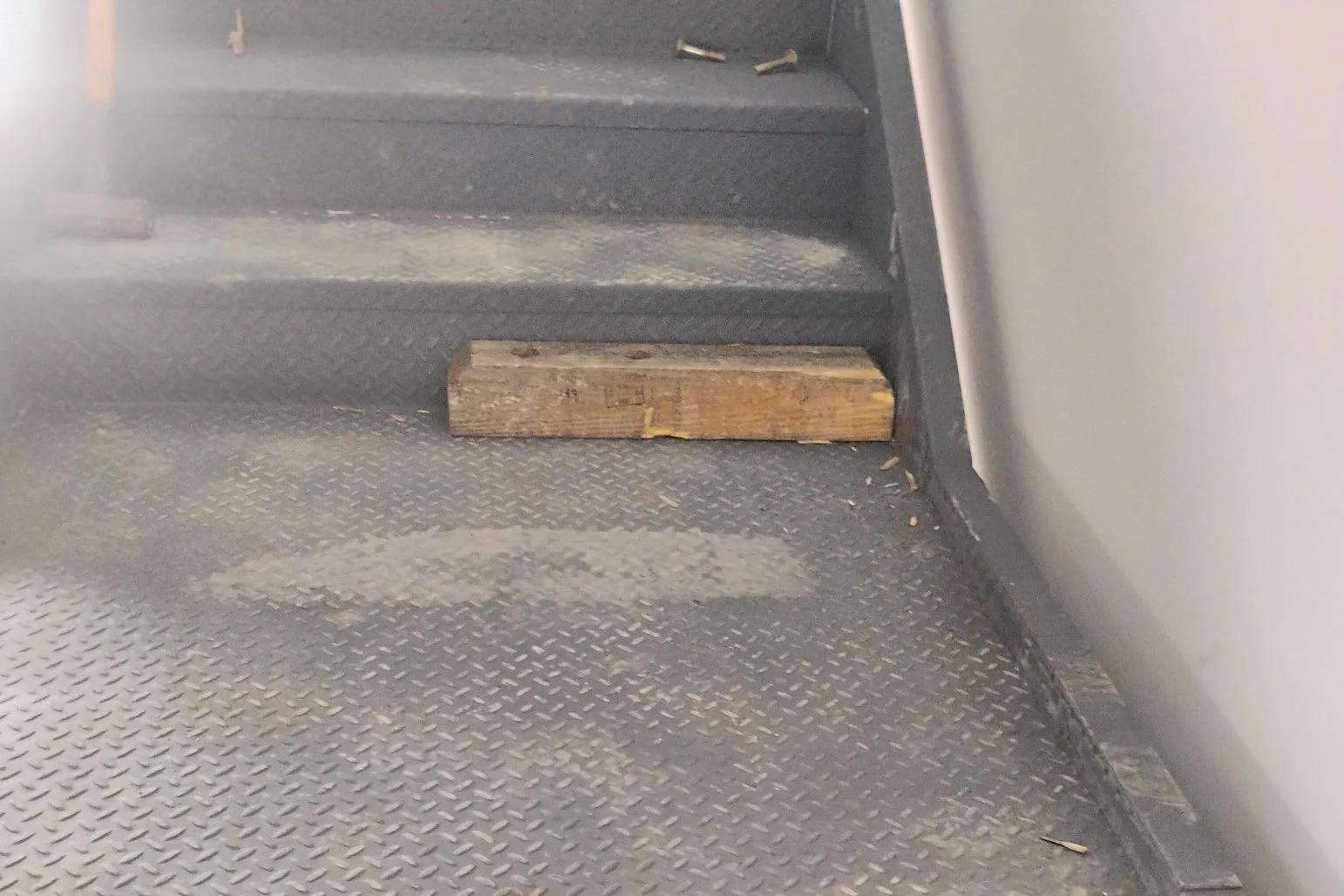 A small wooden block placed on the floor of a vehicle, near the base of a step with a textured rubber surface. There are a few small screws and debris around.