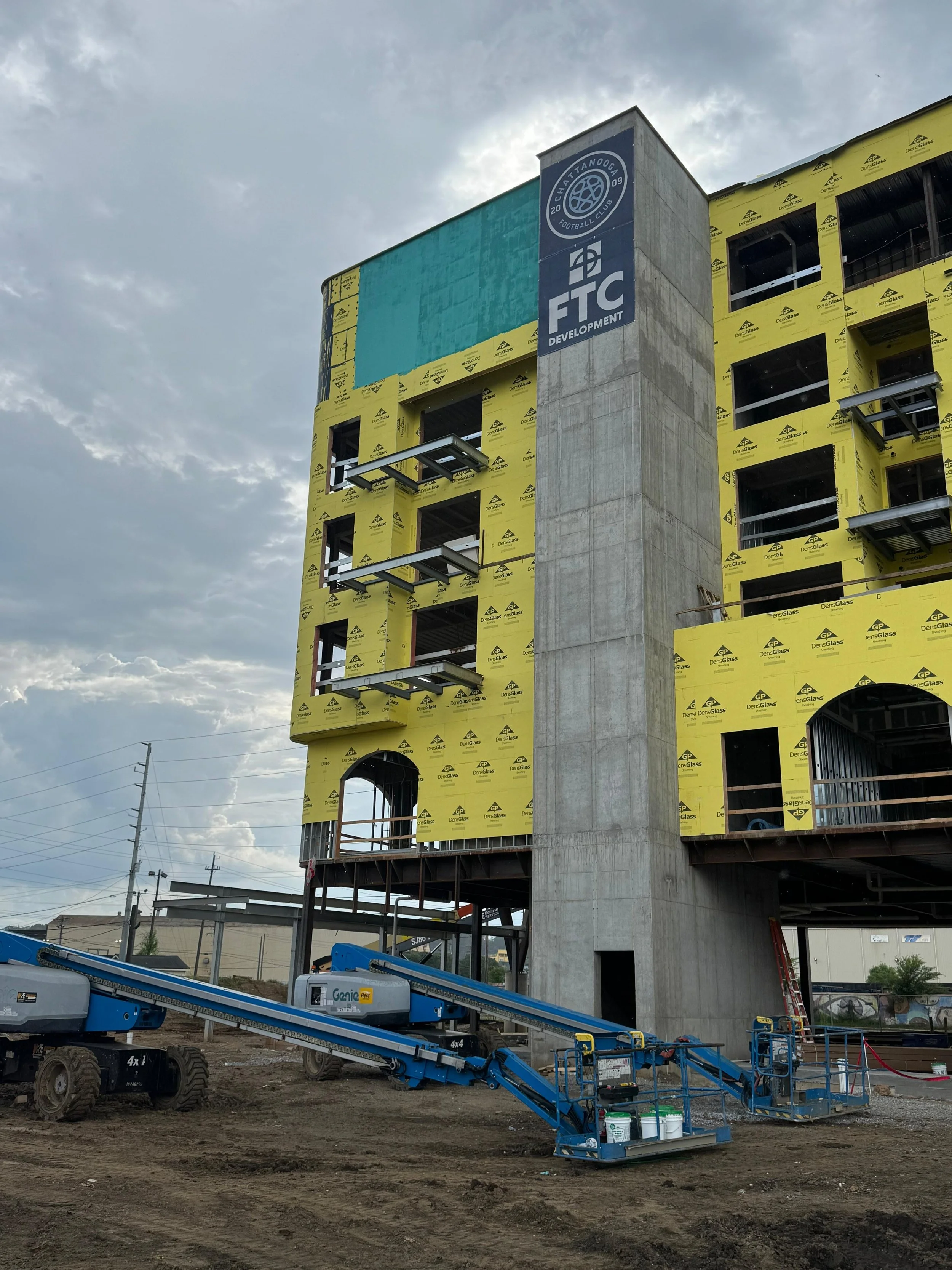 Under construction multi-story building with yellow insulation and gray concrete structure, construction equipment at the ground, cloudy sky.