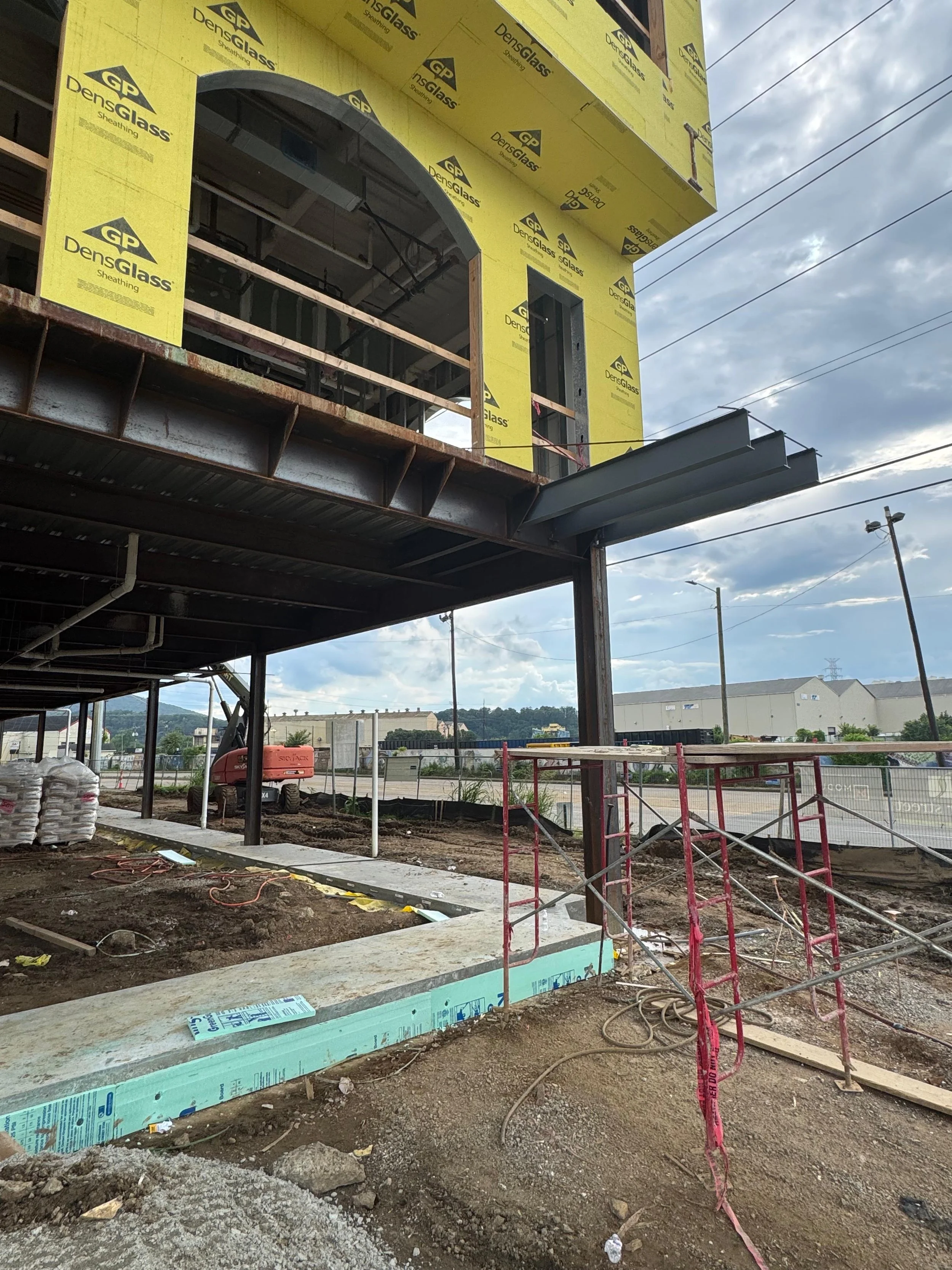 Construction site with partially built building with yellow insulation and metal scaffolding, dirt ground, and cloudy sky.