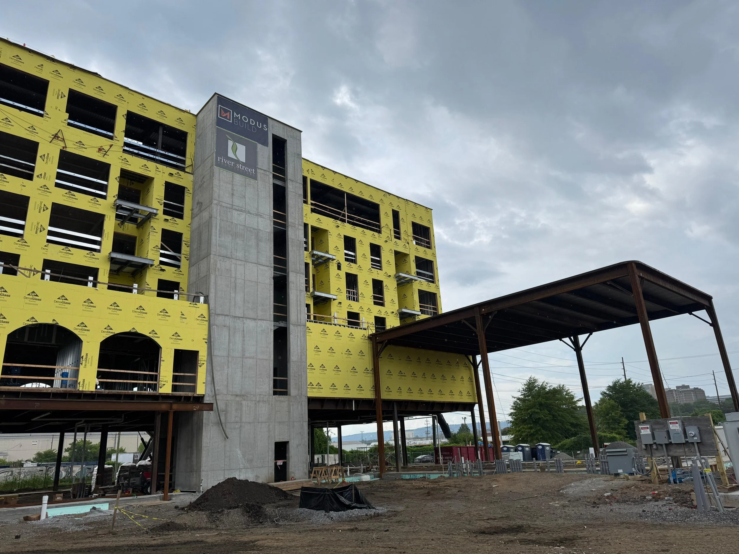 Under construction building with yellow insulation wrap, a concrete elevator shaft, and metal framing for an overhang, with a cloudy sky overhead.