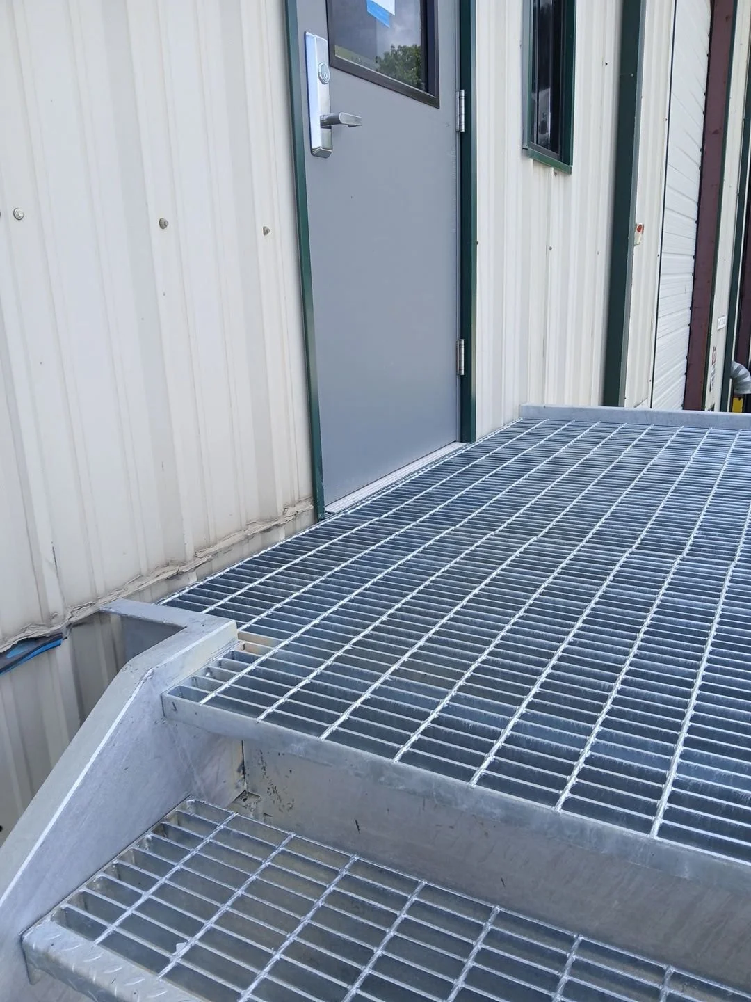 A metal grated ramp outside a building with beige siding and a gray door with a small window and handle.