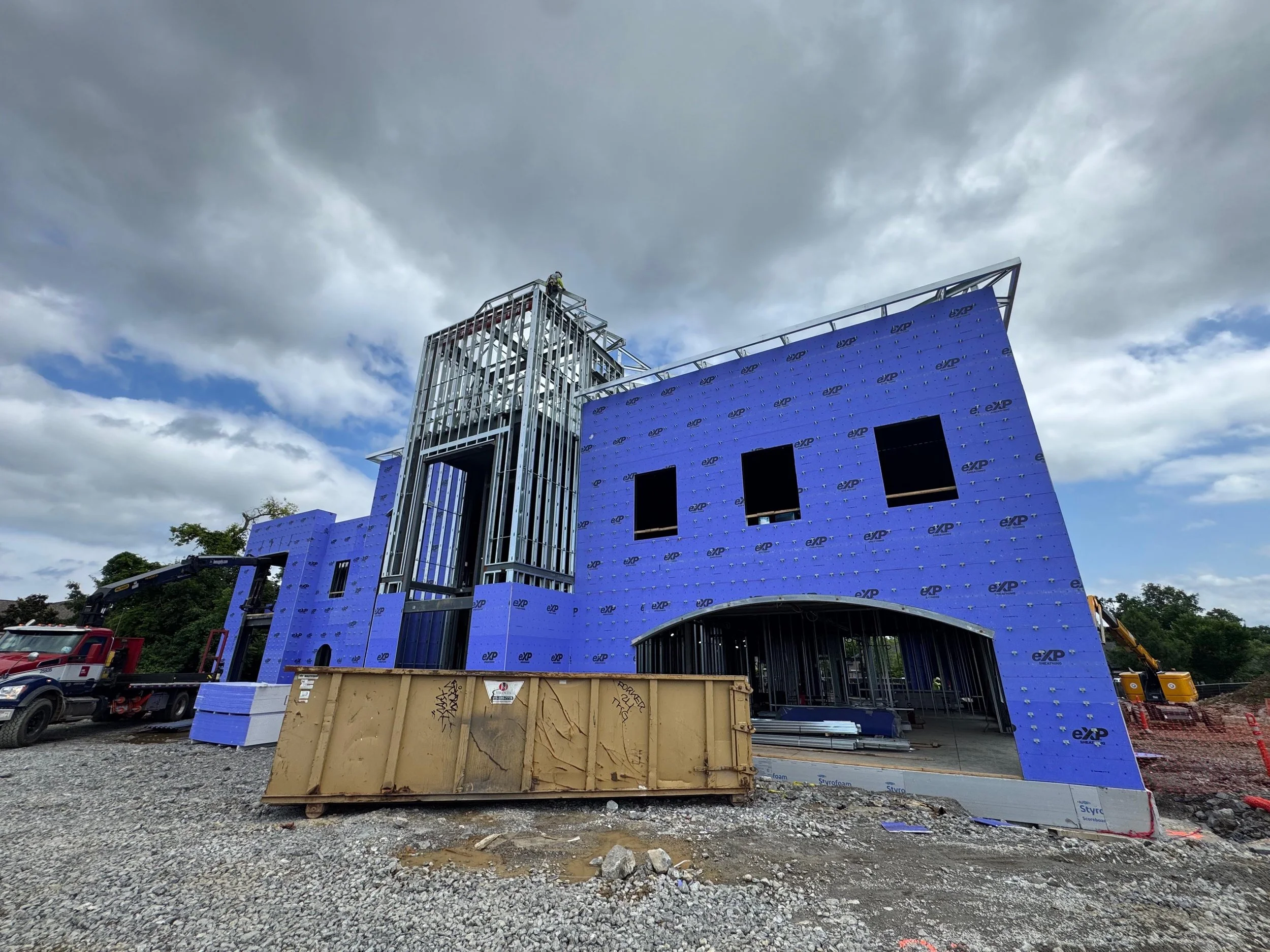 Building under construction with blue insulation wrap, metal framing, and construction equipment, under cloudy sky.