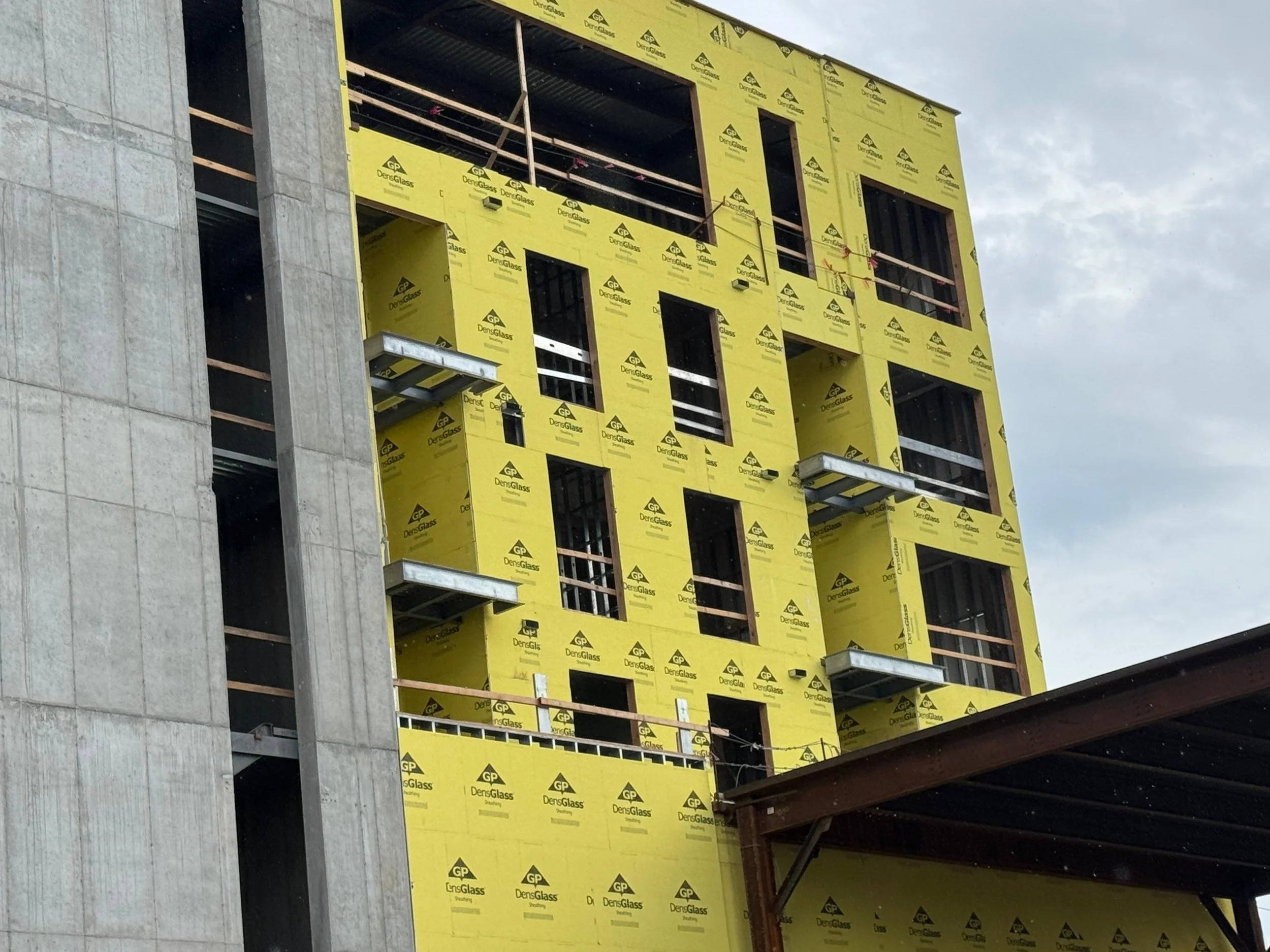 View of a multi-story building under construction with yellow insulation panels and dark window openings, partially covered with scaffolding and construction materials, against a cloudy sky.