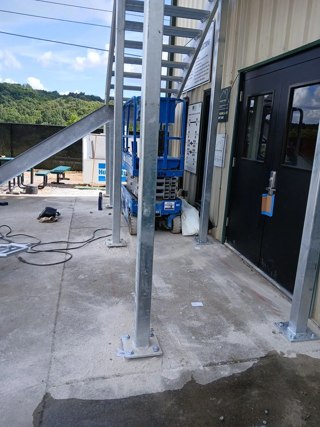 Construction site with metal framework and a blue scissor lift next to a building with signs, above a concrete floor, under a partly cloudy sky.