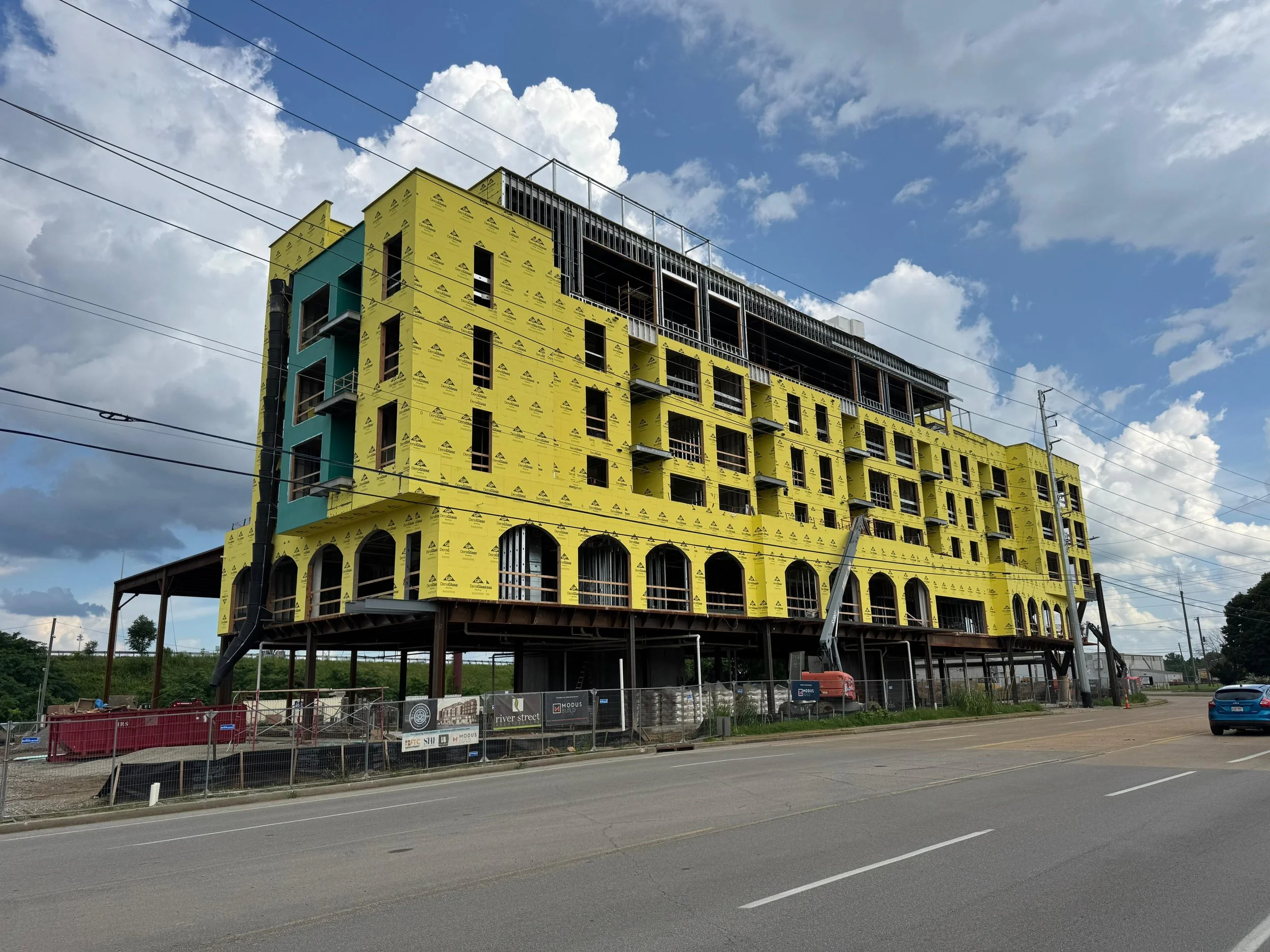 A multi-story building under construction with yellow and teal exterior sheathing, surrounded by construction equipment, fencing, and situated next to a road with cars and power lines, under a partly cloudy sky.