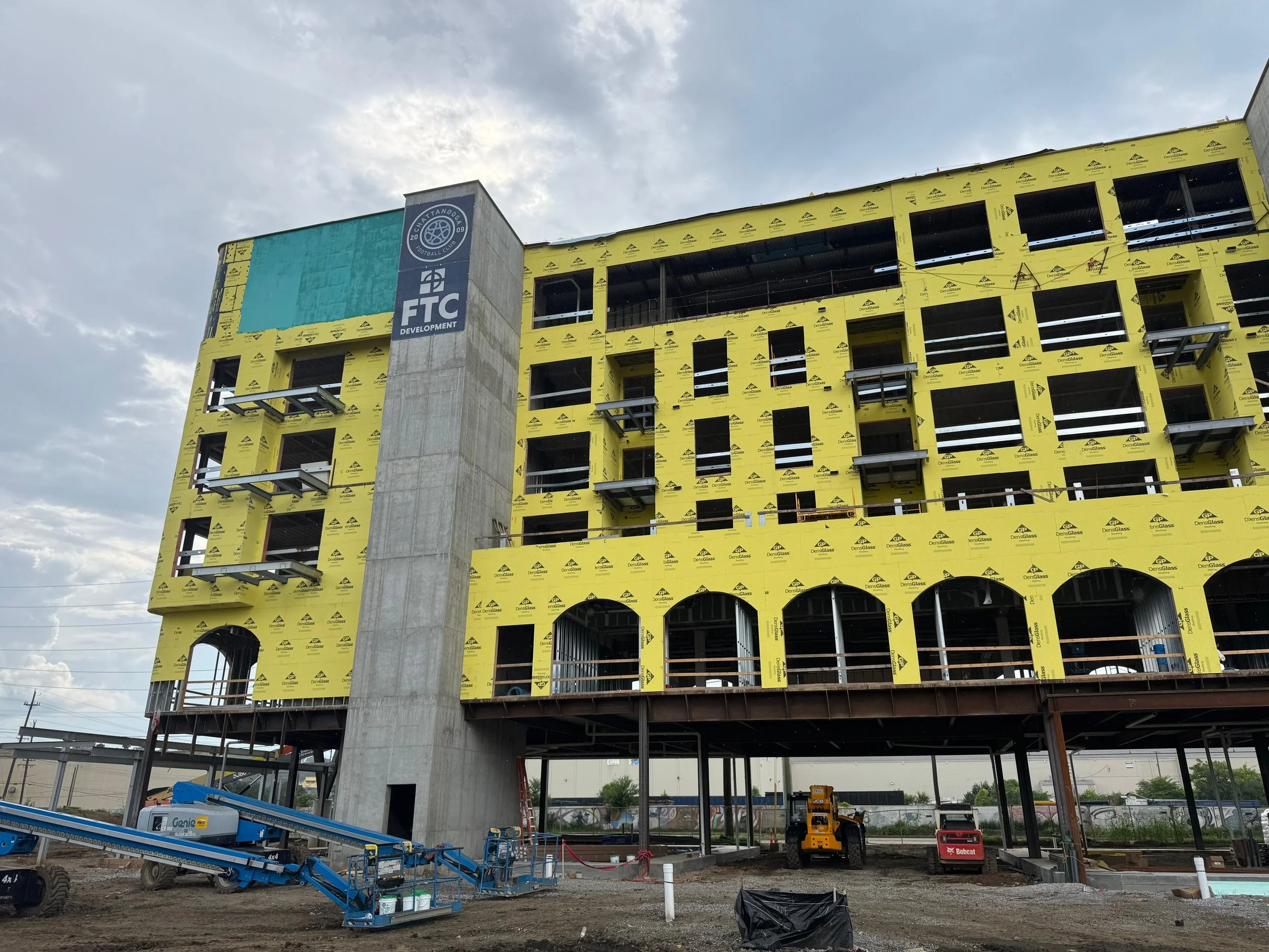 Under construction building with yellow sheathing, multiple floors, balconies, and a large blue and gray sign that reads 'FC Development.' Construction equipment is on the ground level near the building.
