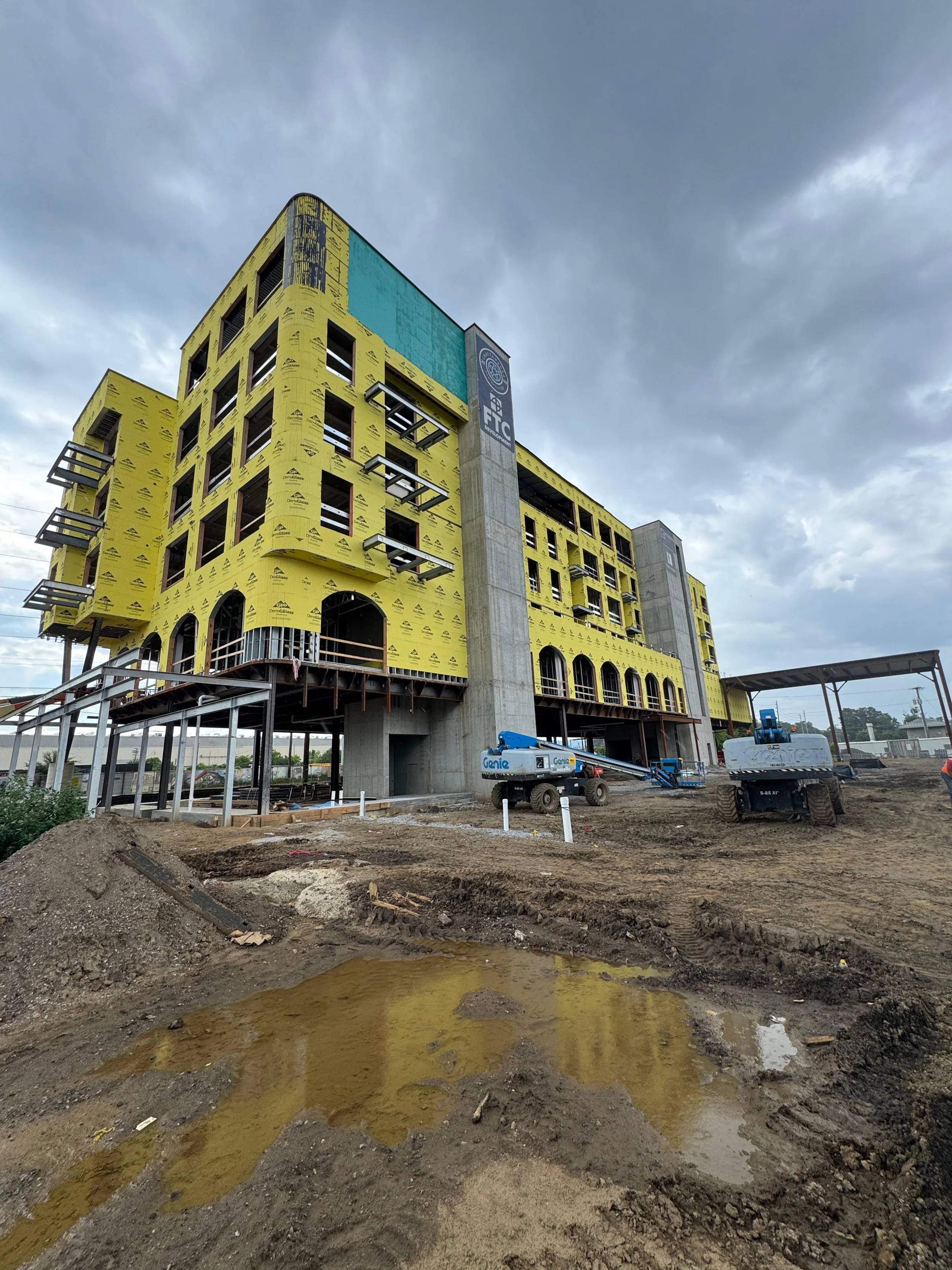 Construction site with a partially built yellow multi-story building, construction equipment, and cloudy sky.