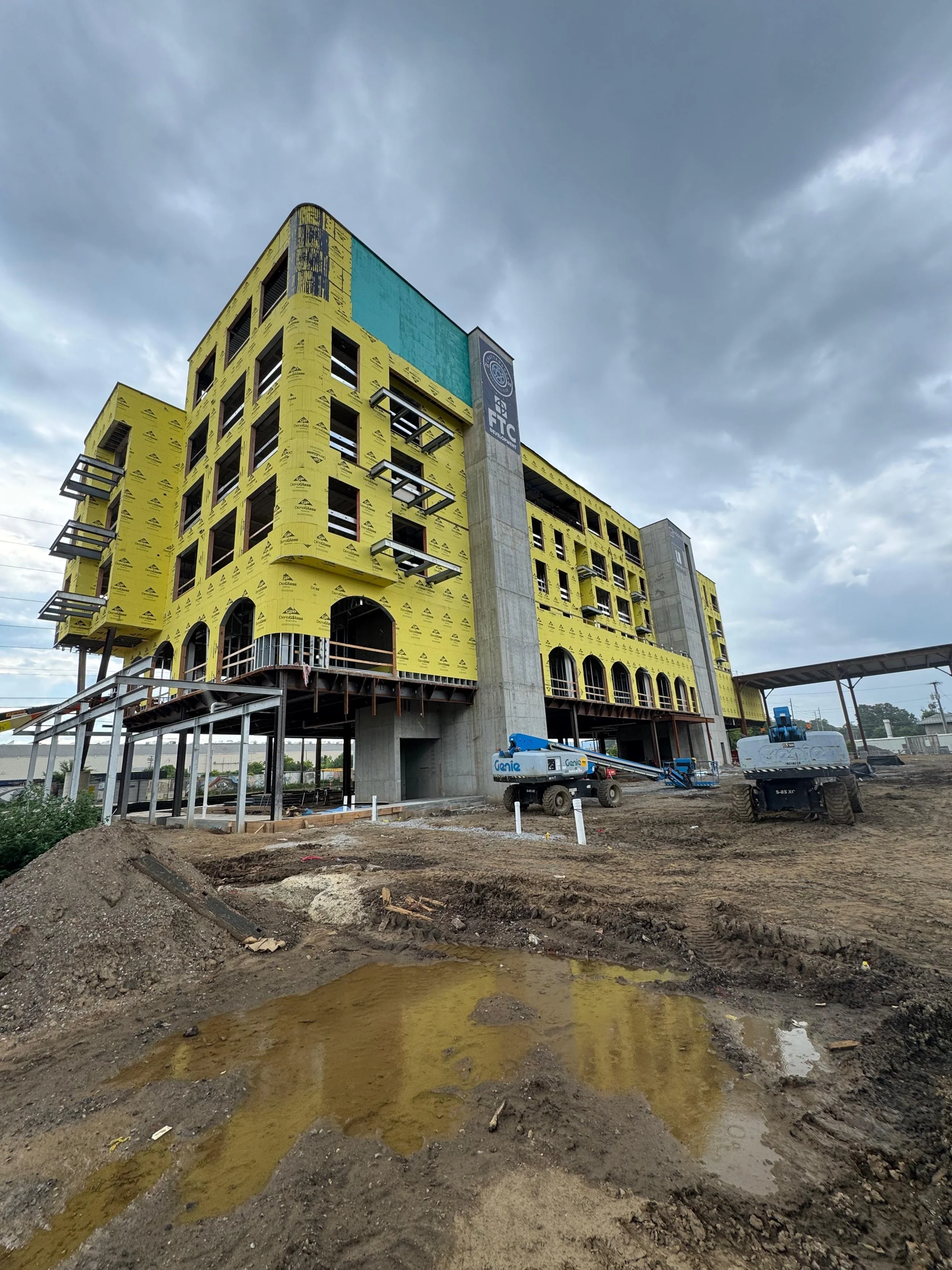 Under construction multi-story building with yellow insulation panels, construction equipment, muddy ground with puddles, and cloudy sky.