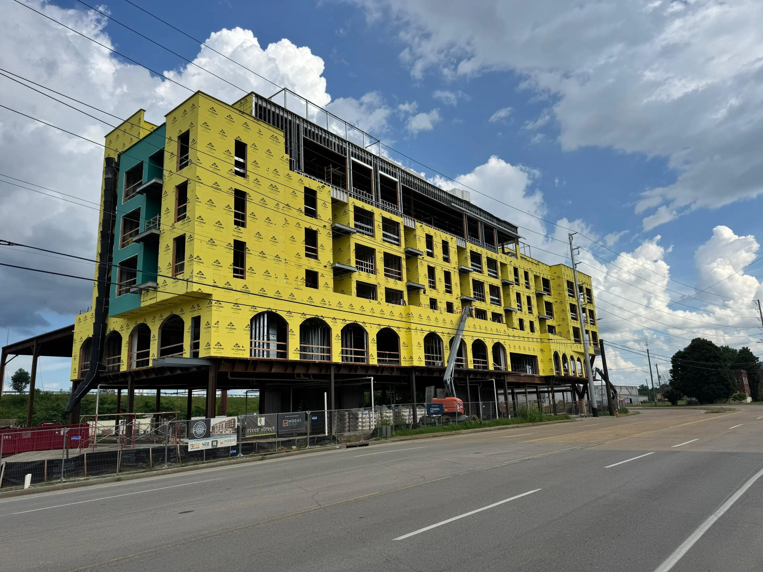 A multi-story building under construction with a yellow exterior wrap, scaffolding, and construction equipment, near a road with power lines and cloudy sky.