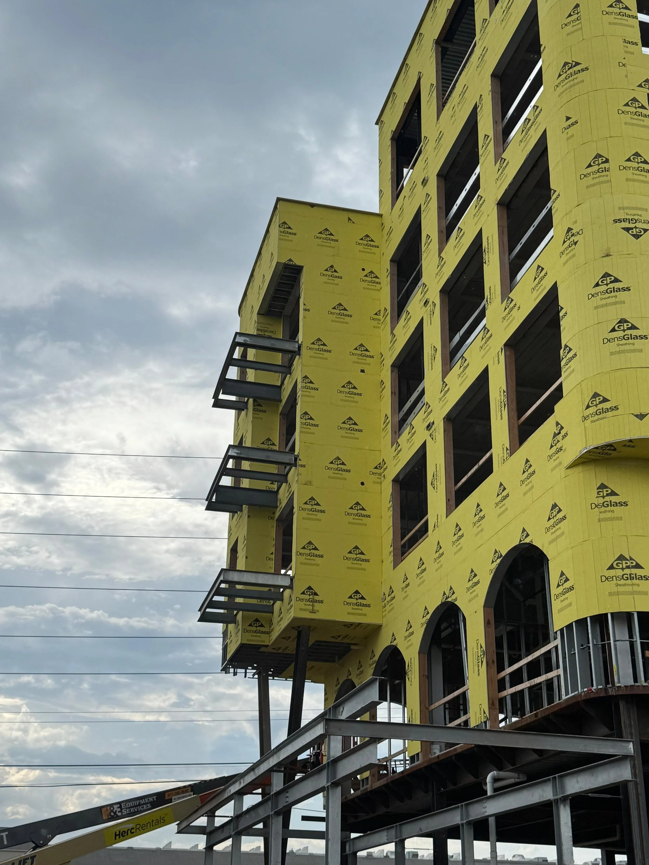 Under-construction building wrapped in yellow insulation material with window openings and small balconies, against a cloudy sky.
