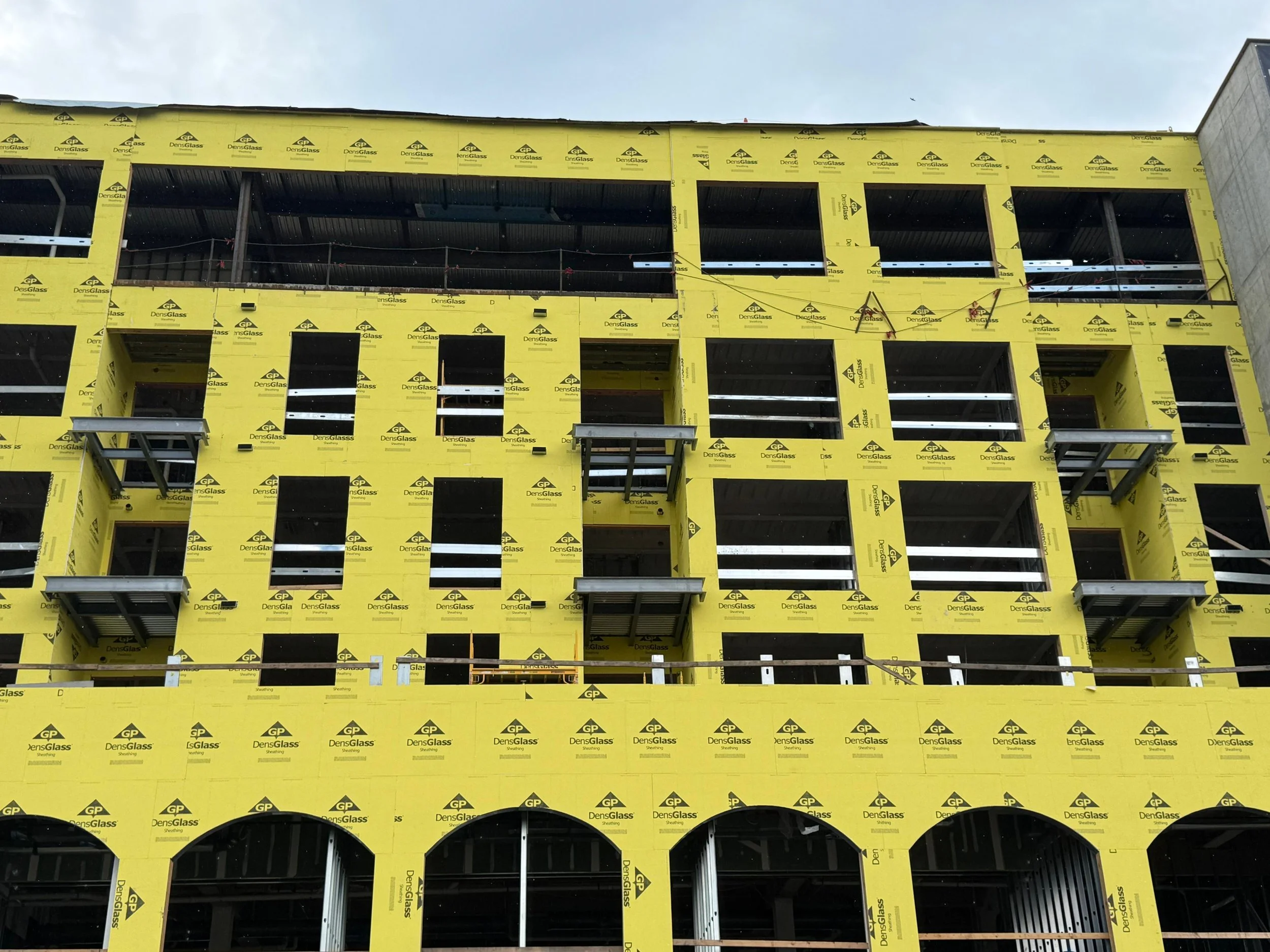 Building under construction with yellow insulation wrap and open window spaces, scaffolding, and construction barriers.