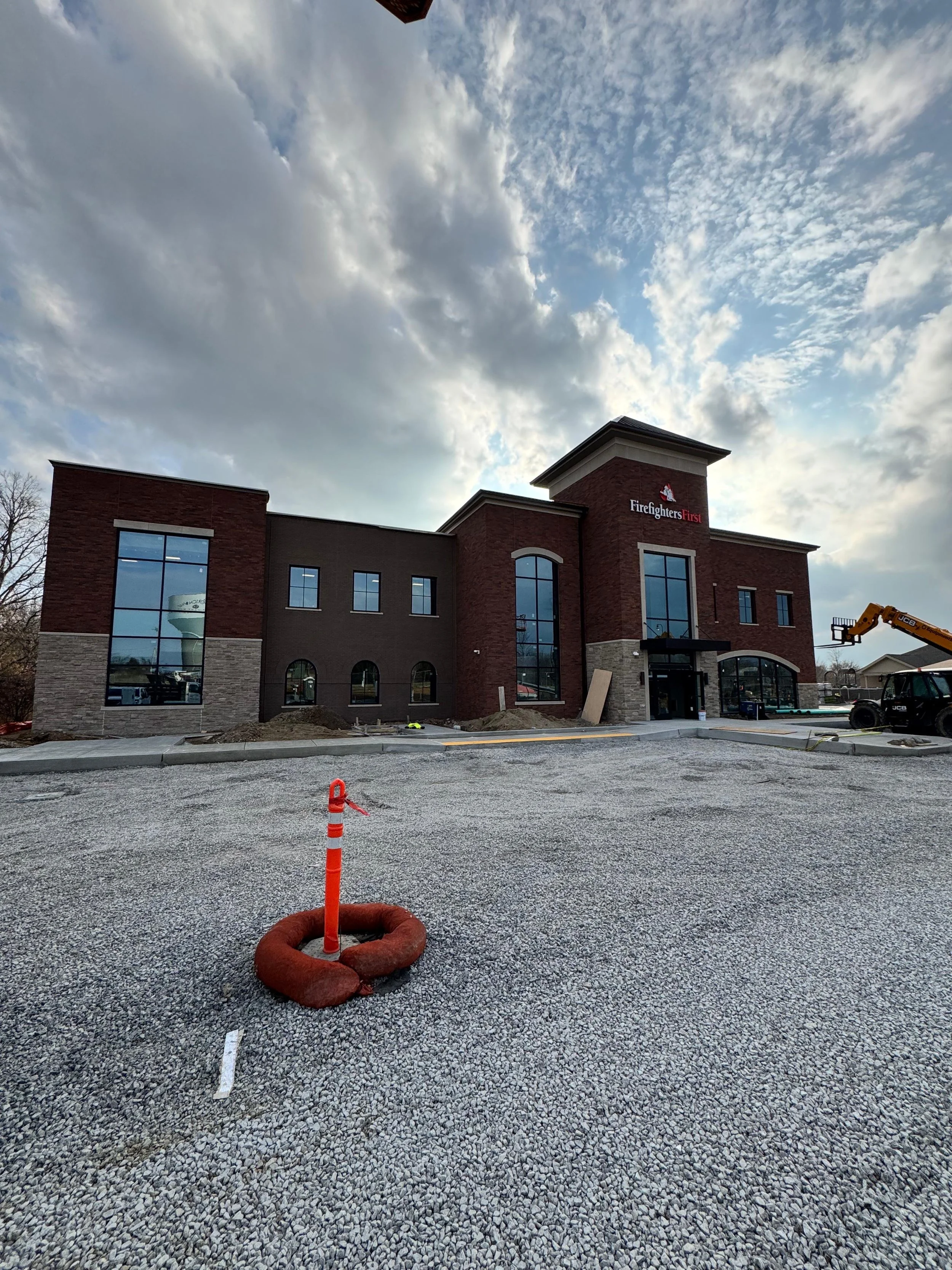 Construction site in front of a two-story brick building with glass windows, labeled 'Firefighters First,' under a cloudy sky with some blue patches.