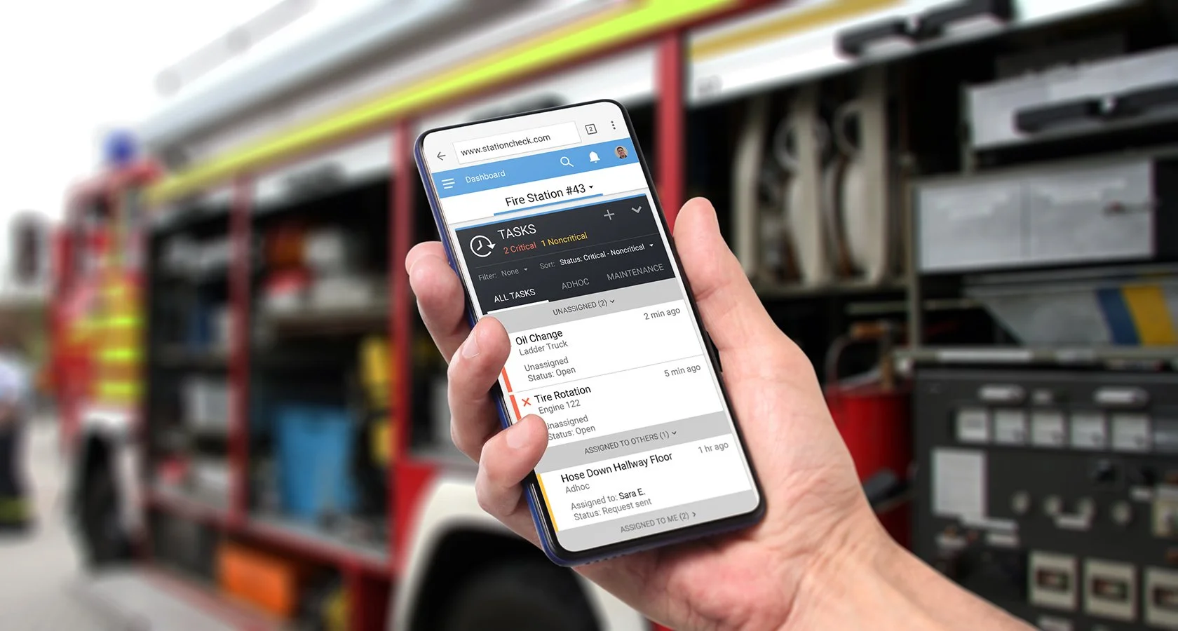 Hand holding a mobile device with a firehouse equipment management app displayed. Firetruck in background