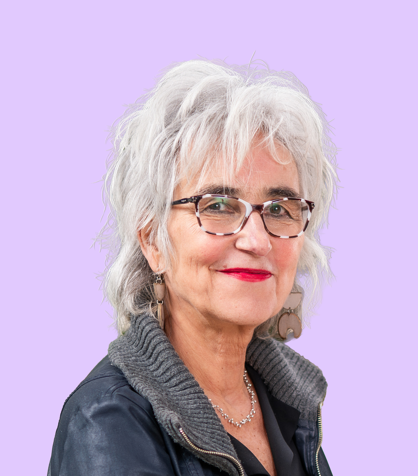 Close-up of a mature woman with silver hair, wearing glasses, a black top, a gray knit scarf, and a leather jacket, against a light purple background.