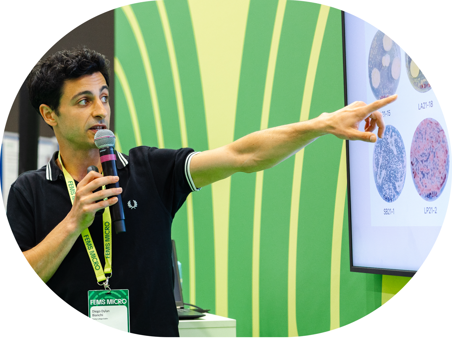A man holding a microphone and pointing at a large screen displaying microscopic slides of cells during a presentation at FEMS Micro conference.