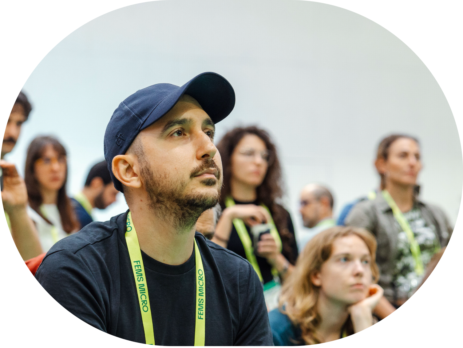 A man wearing a blue baseball cap and black T-shirt listening attentively at a conference or seminar, with several other attendees in the background.