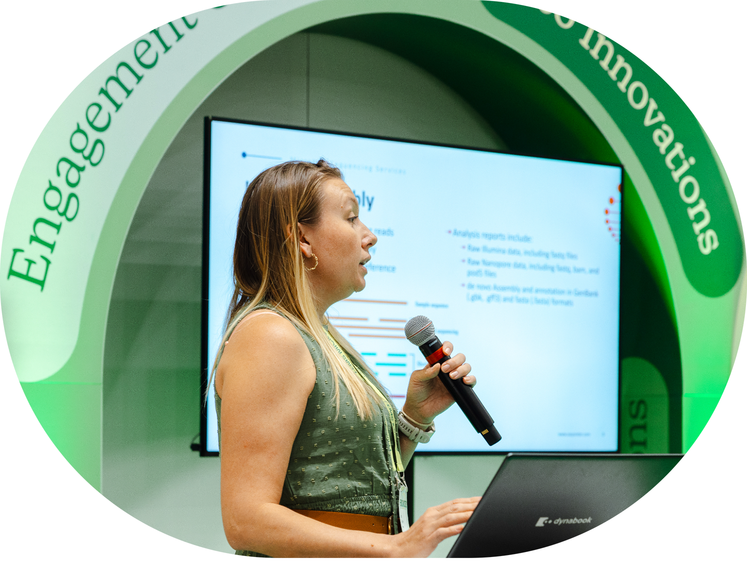 A woman with blonde hair wearing a green sleeveless dress speaking into a handheld microphone at a presentation. She is standing in front of a large screen displaying a slide with text and graphs, and a green archway with the words "Engagement" and "