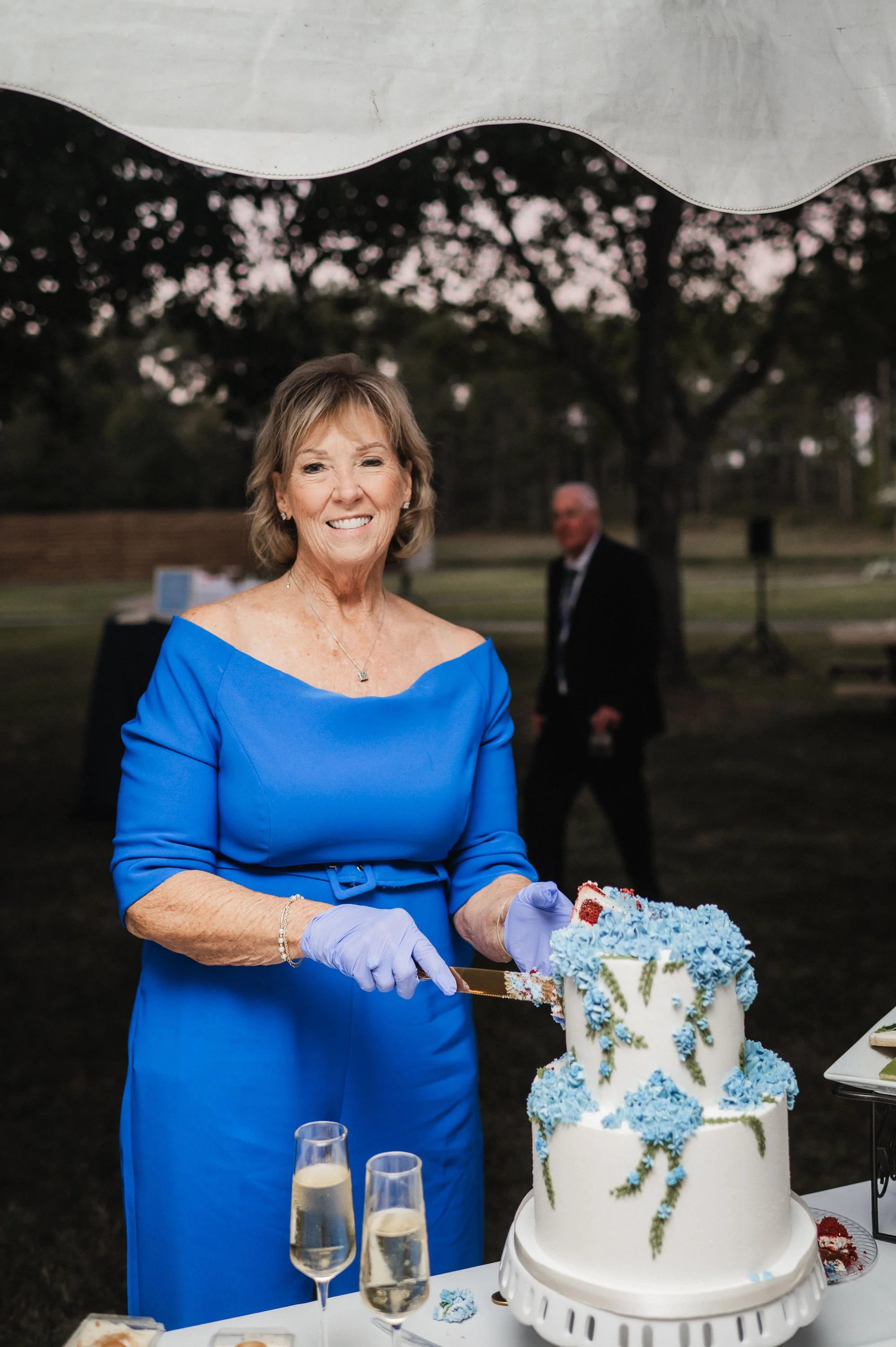A smiling woman in a blue dress, wearing lilac gloves, is holding a knife and cutting a white wedding cake decorated with blue flowers. There are two glasses of champagne on the table. In the background, a man in a suit is blurred and standing outdoors near trees at sunset.