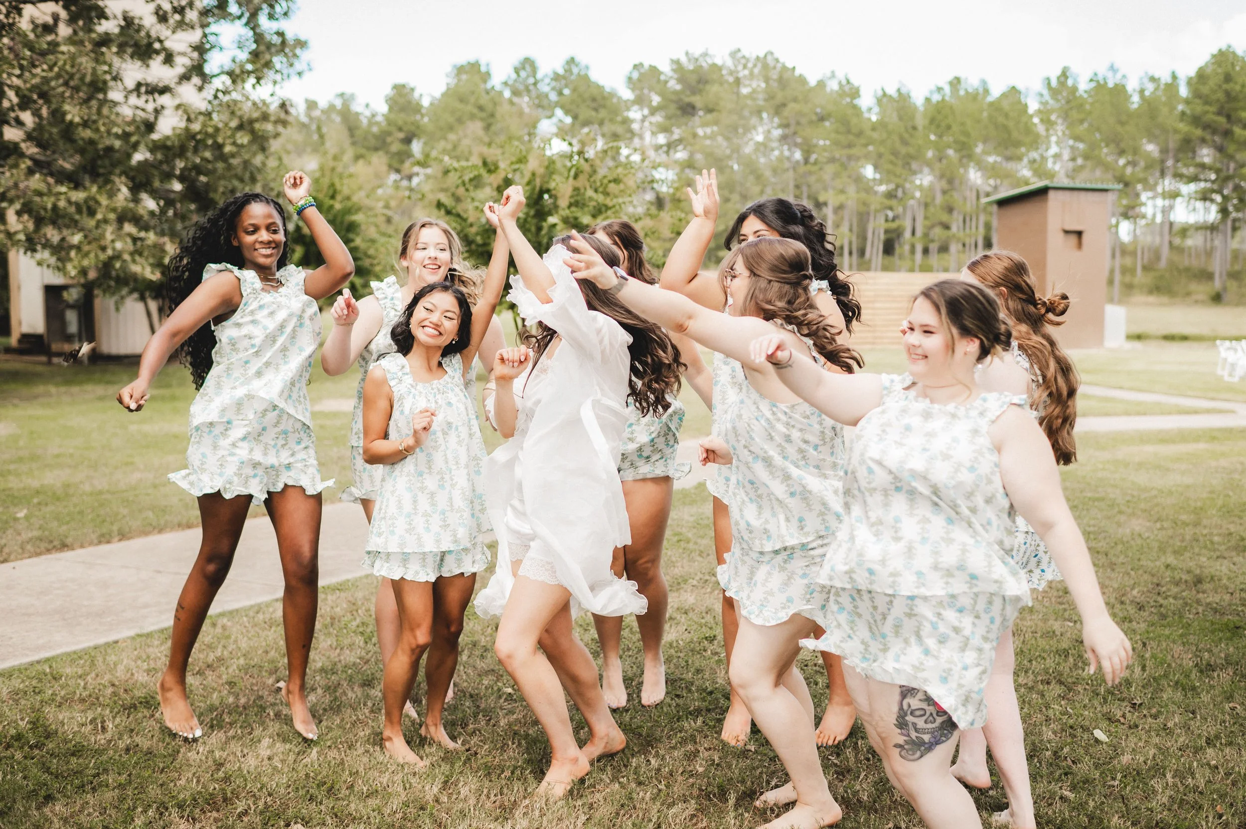 Group of women and girls wearing matching white sleepwear with blue floral prints, enjoying a dance party outdoors on the grass with trees and a building in the background.