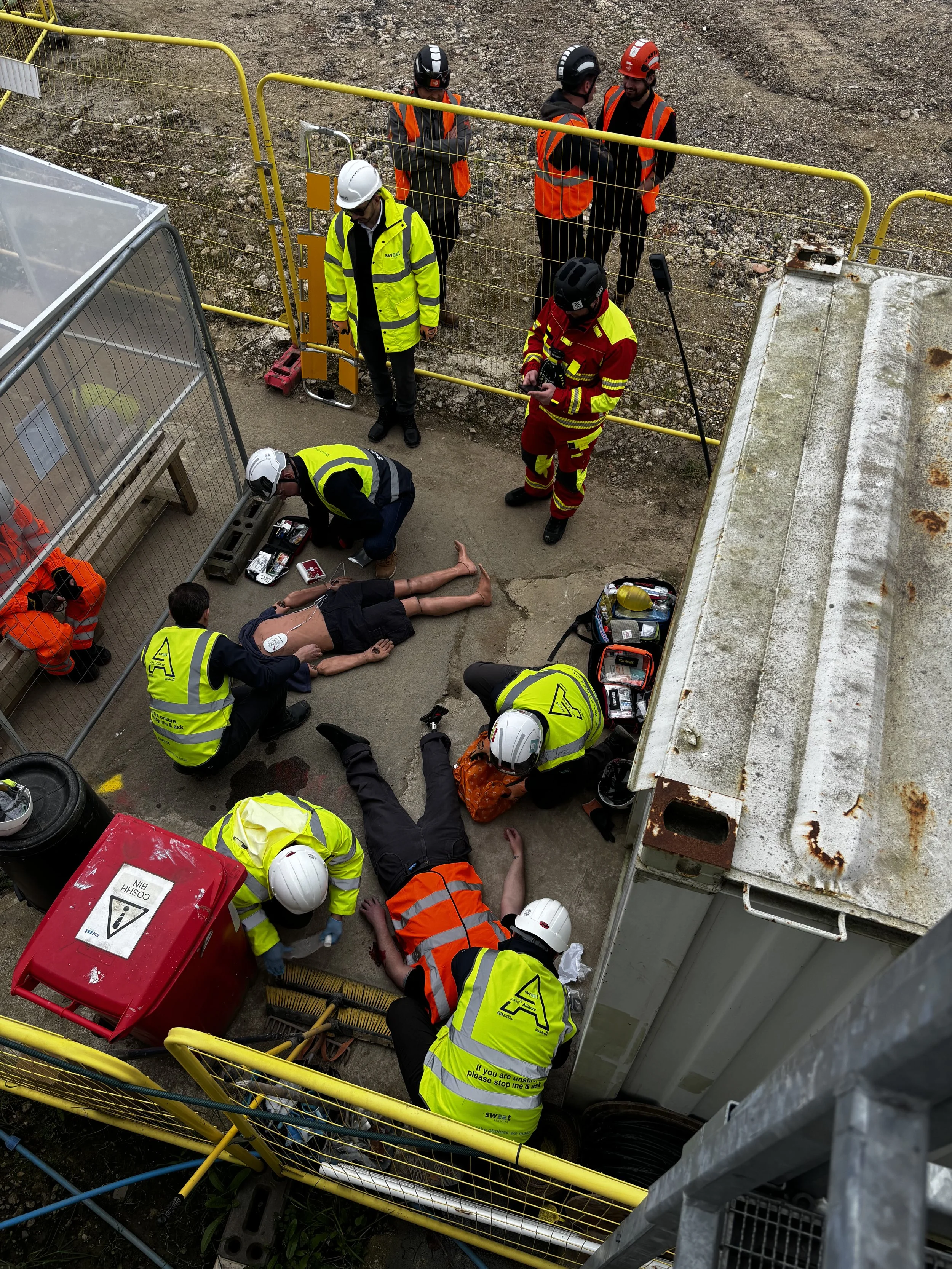 Group of emergency responders and workers attending to a patient on a stretcher inside a warehouse or industrial facility. Some individuals are wearing orange uniforms with "radius" and "PARAMEDIC" labels, while others are in casual and safety gear.