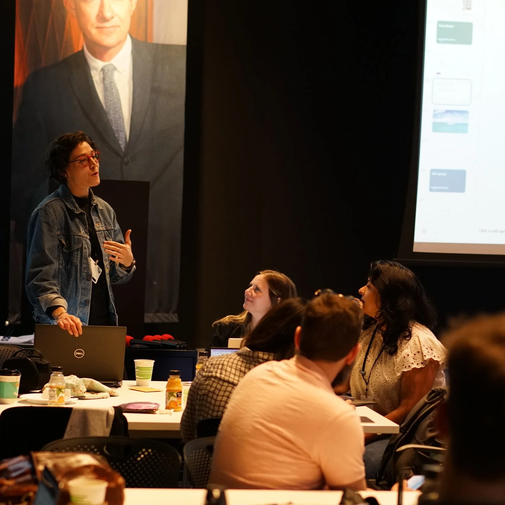 Woman in jean jacket speaks with group of people sitting at tables