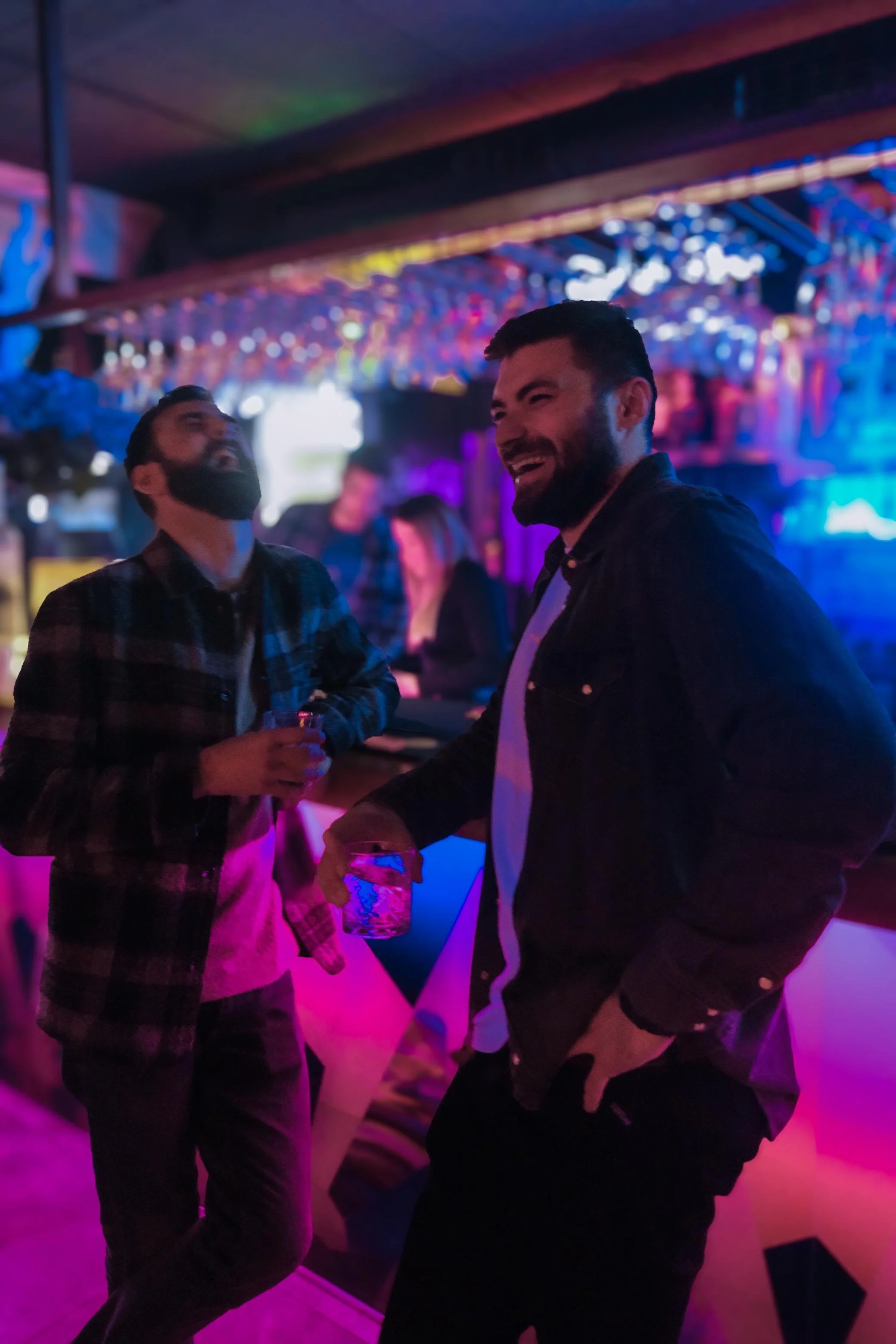 Two men laughing and socializing at a bar or nightclub with colorful, neon lighting.