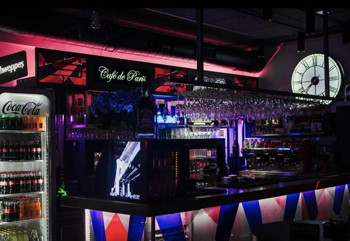 Bar area with neon lights, a Coca-Cola refrigerator, hanging wine glasses, and a large clock in a dimly lit nightclub or bar