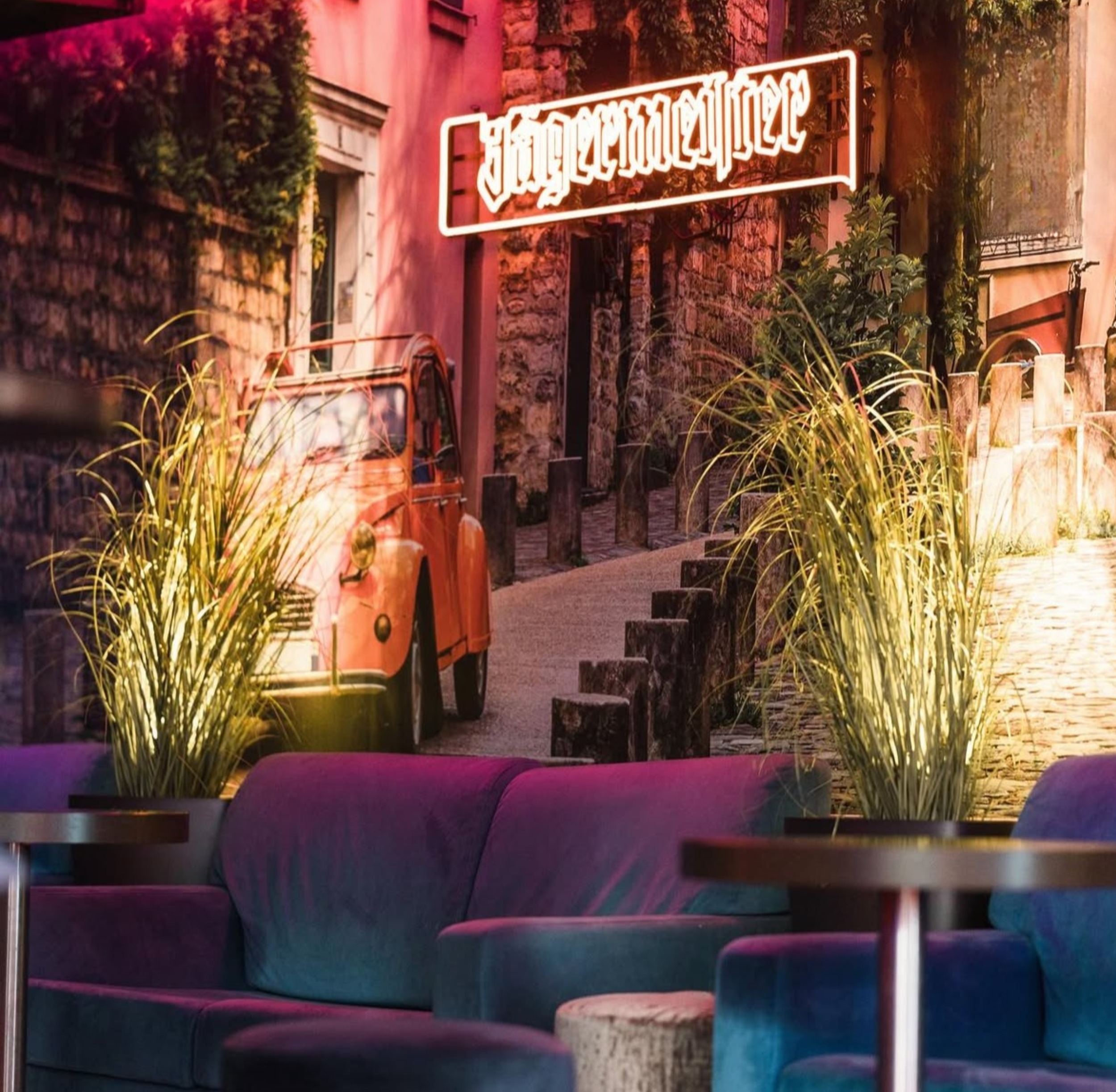 Interior with purple sofa, small round tables, indoor plants, and a large mural of a street scene with a vintage orange car, a stone building, and a neon sign reading 'Hoppadaria' in a Old Town setting.