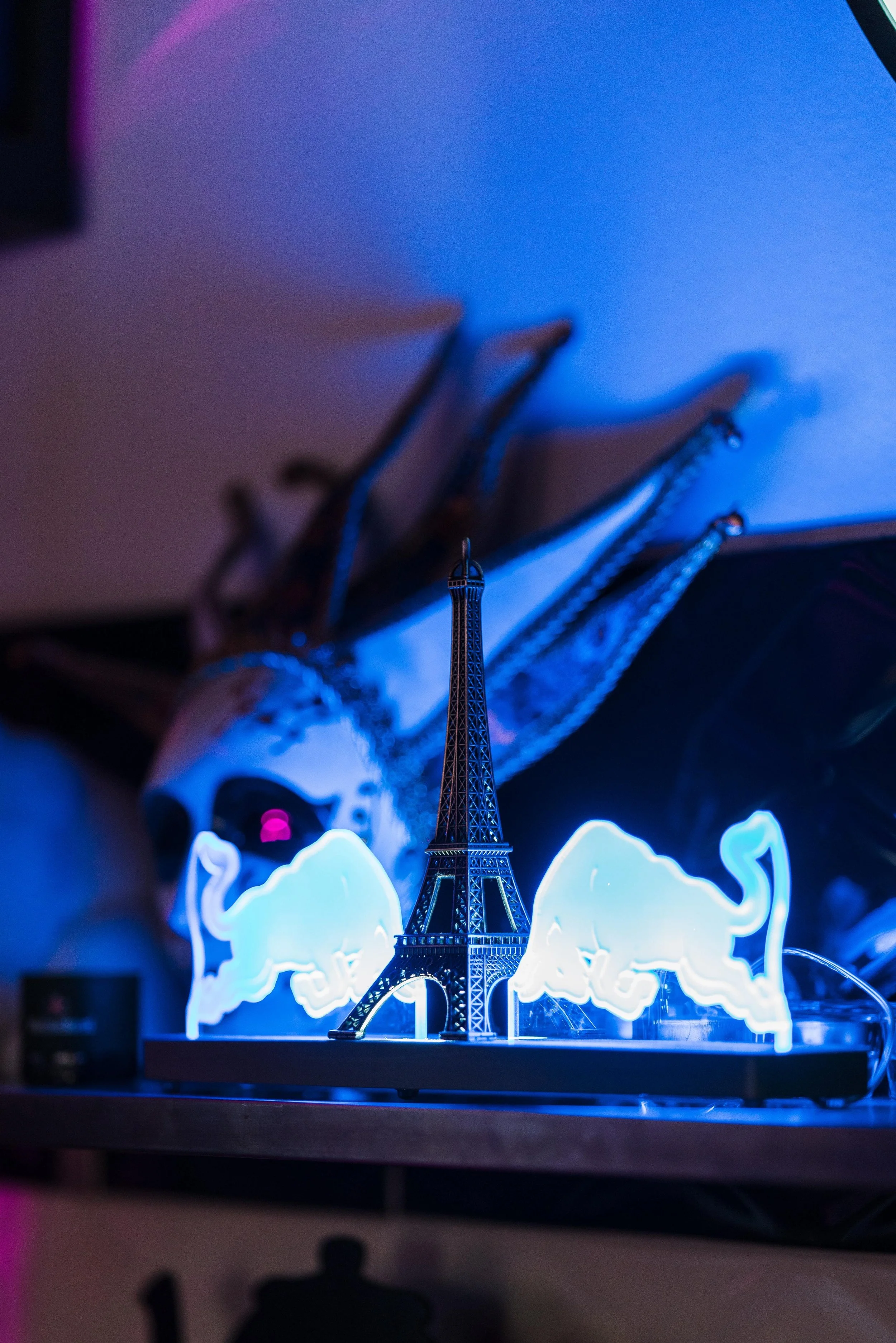 A miniature Eiffel Tower statue illuminated by neon lights, with two neon bull logos on either side, placed on a black base on a wooden surface.
