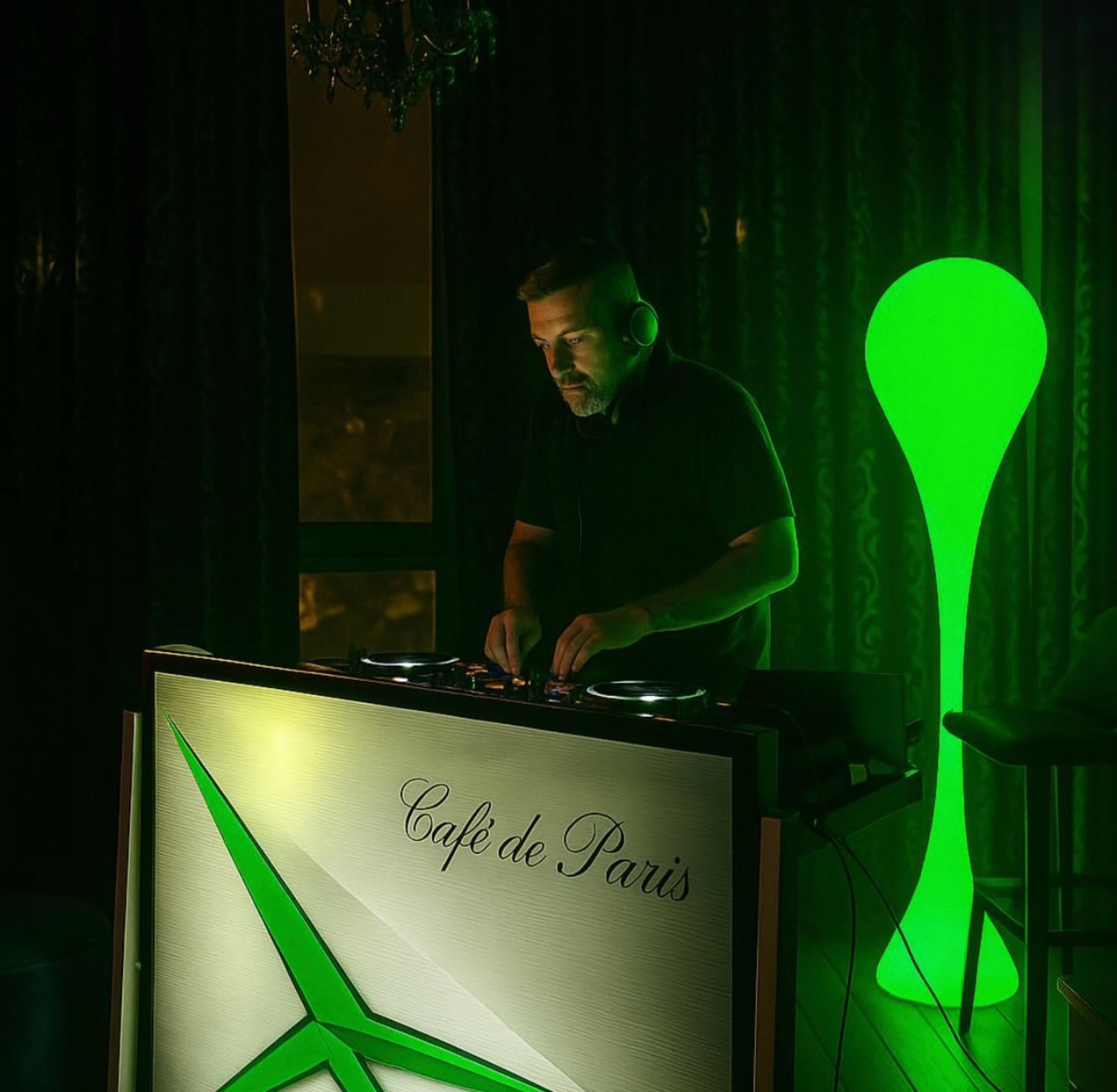 A DJ in a dark room with green lighting, wearing headphones, standing behind a DJ booth that reads 'Café de Paris' with a green and white logo.