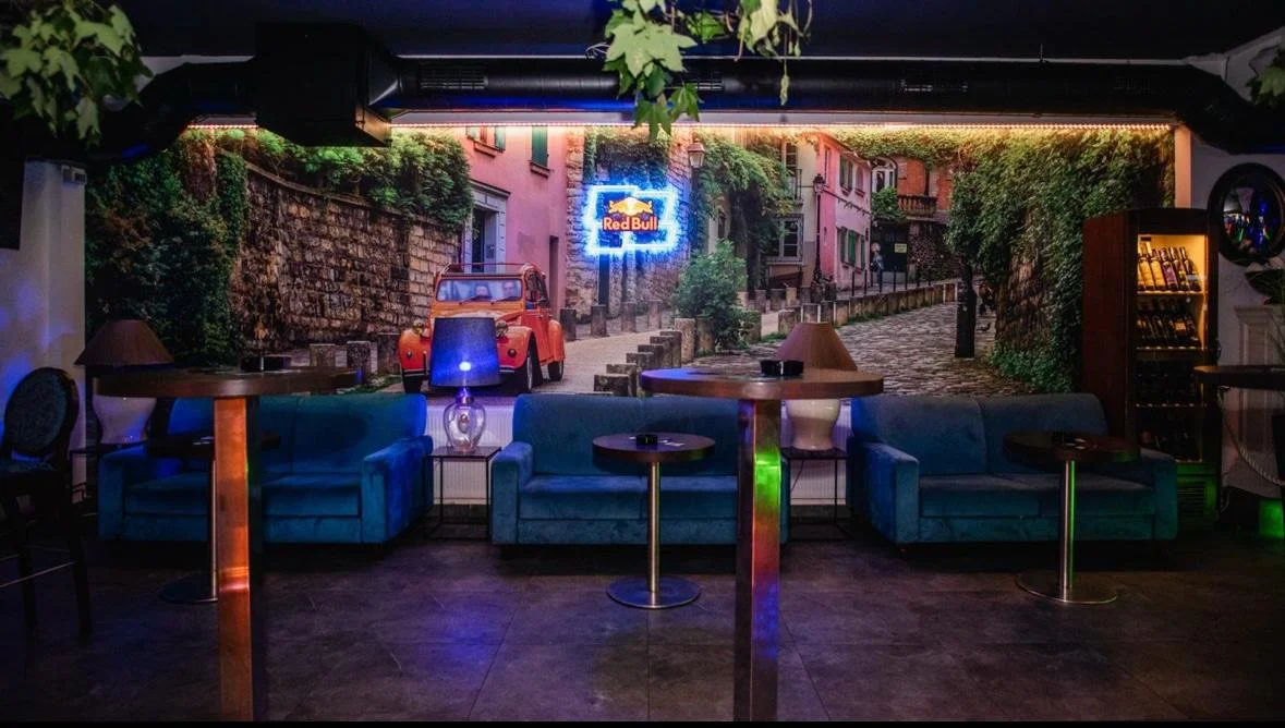 Indoor lounge area with three blue couches, small tables, and a wall mural of a street scene with a vintage red car, plants, and a neon Red Bull sign.