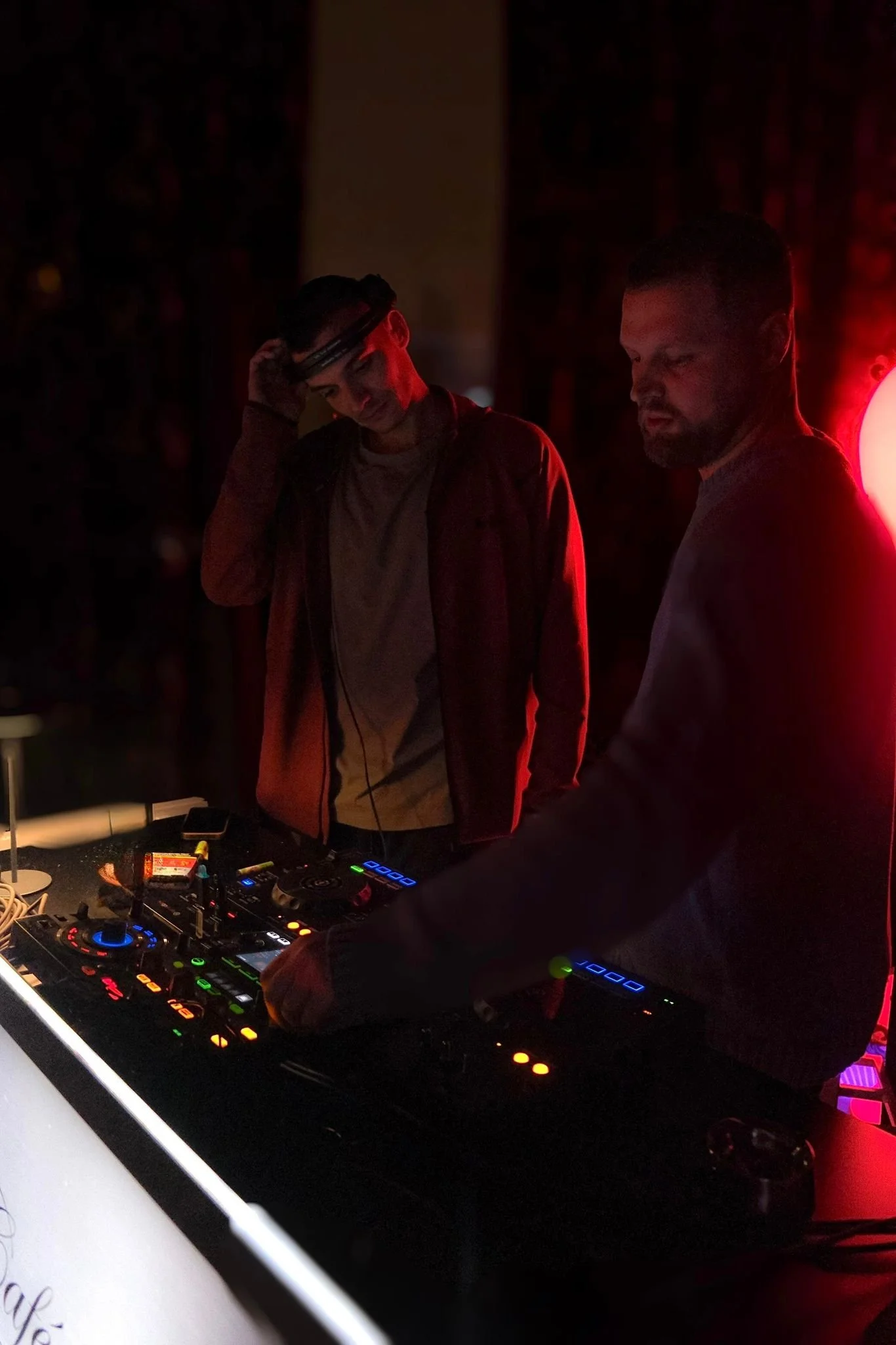 Two men working as DJs at a dimly lit event, using electronic DJ equipment, with red lighting creating a moody atmosphere.