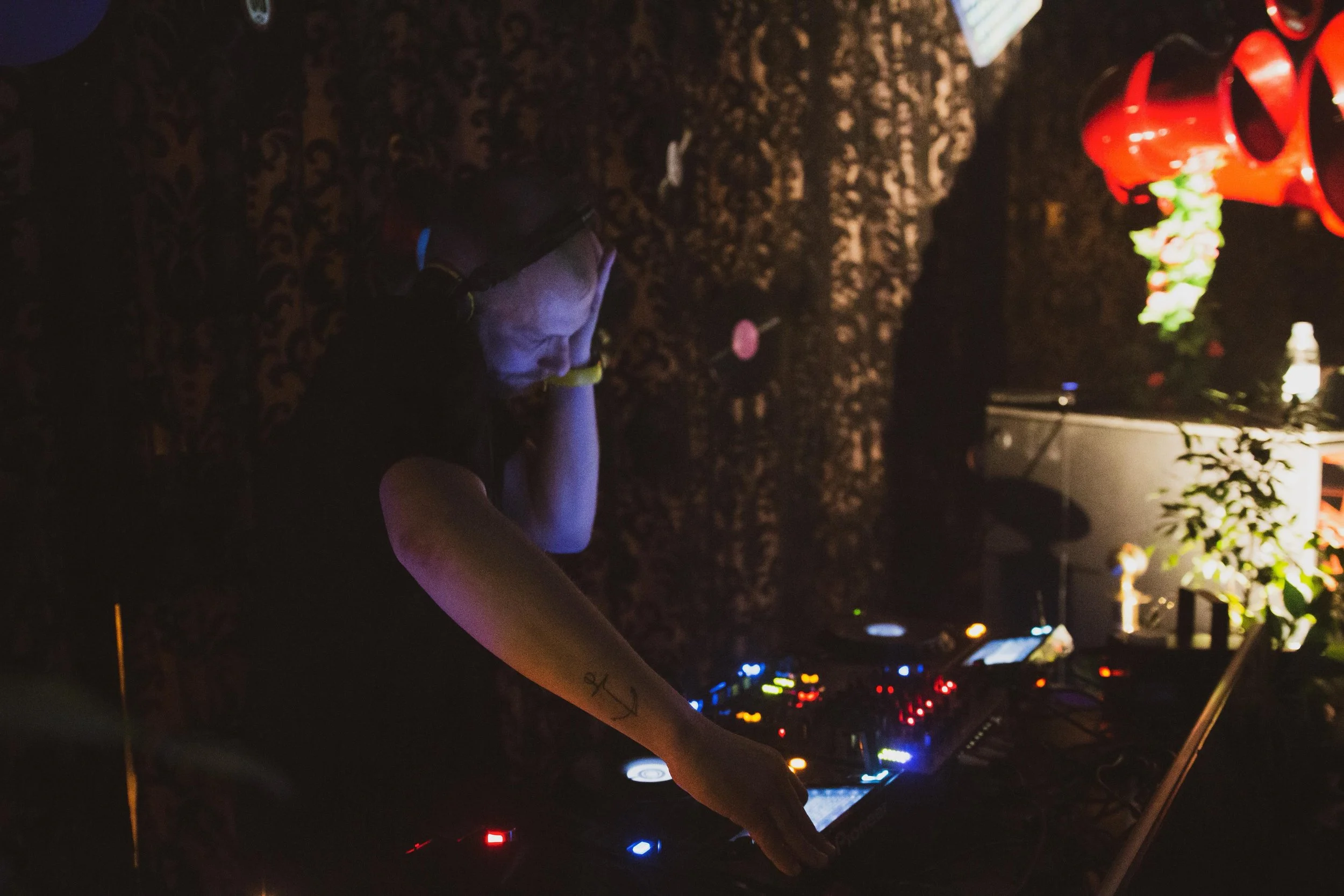 A DJ wearing headphones is mixing music at a dimly lit party with colorful lights and decorative plants.