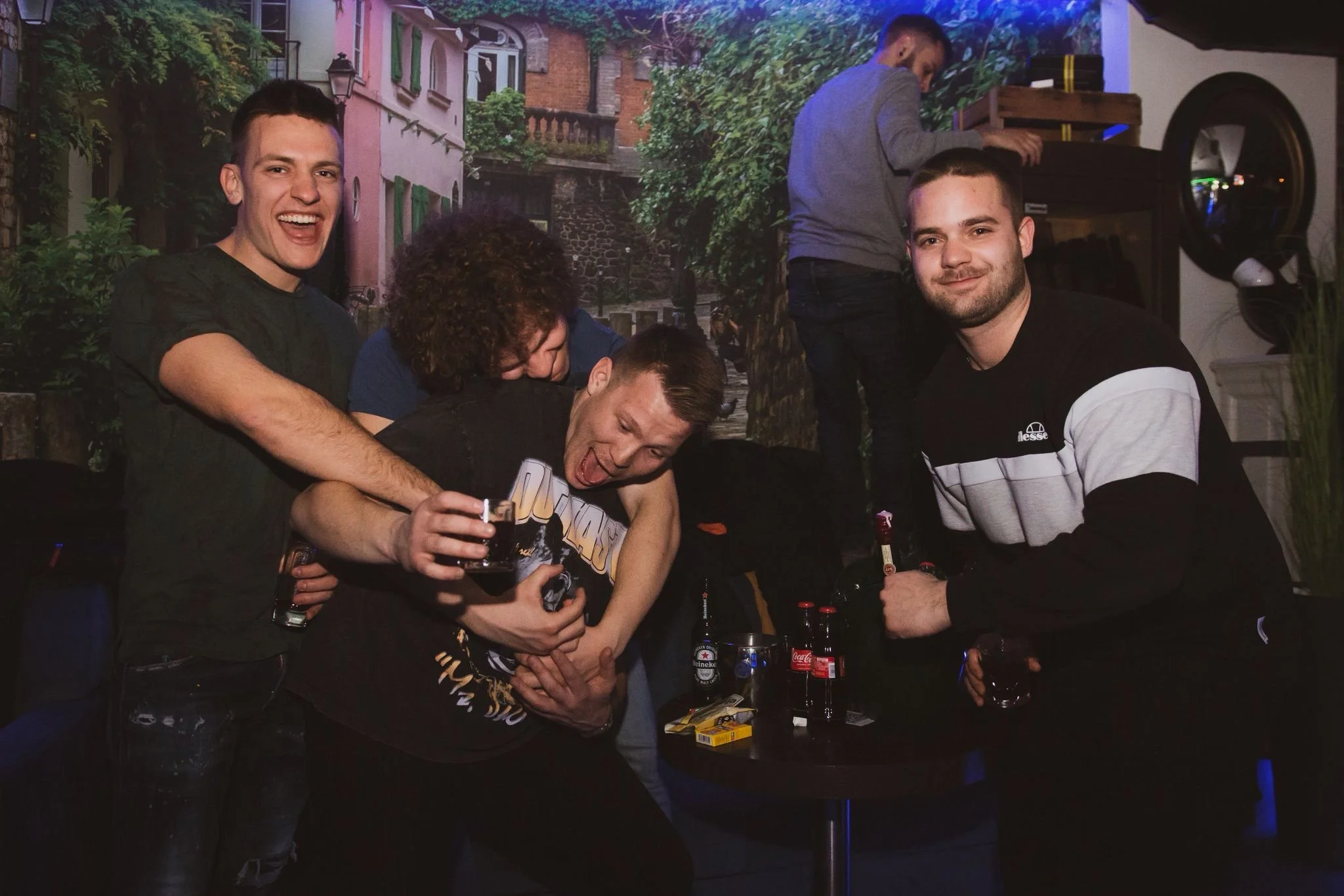 Four young men having fun at a bar, some holding drinks, with a background mural of a street scene with trees and buildings.
